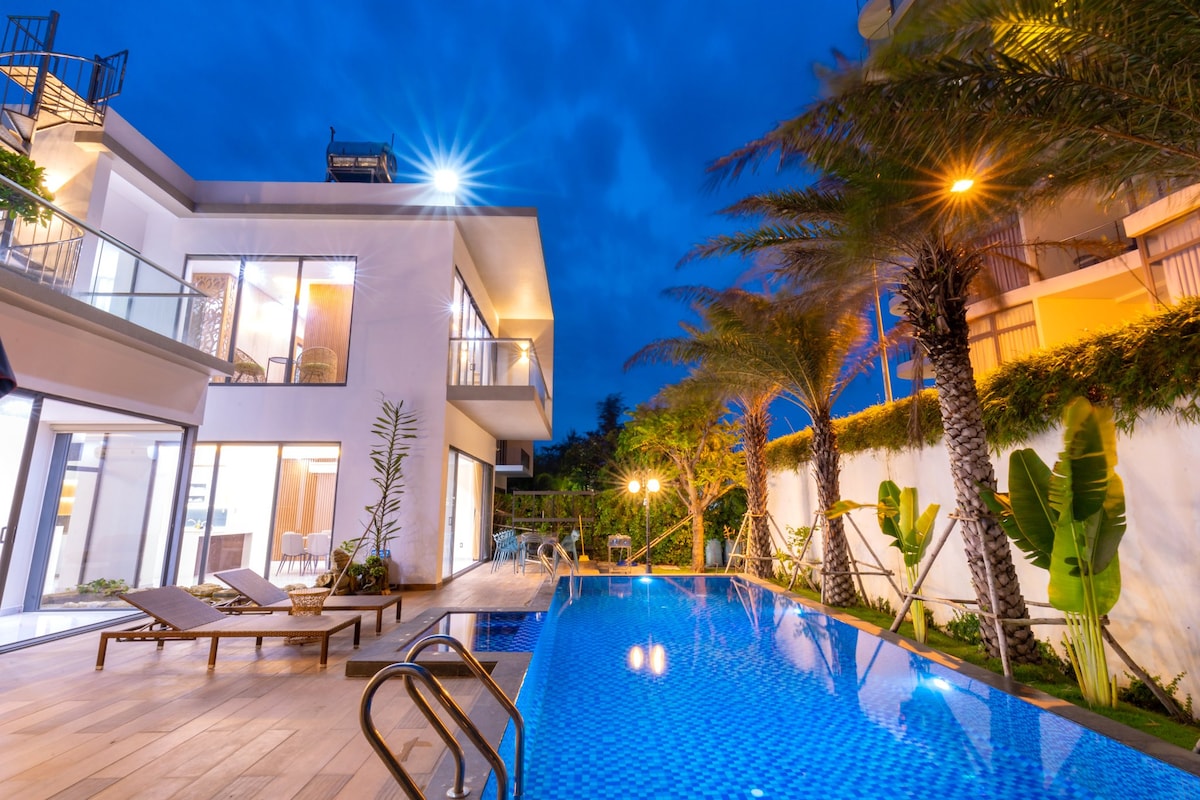 The villa is illuminated by soft outdoor lighting, showcasing a clear blue swimming pool surrounded by lush greenery. Loungers are positioned elegantly near the pool, and large glass doors reveal the spacious interior, enhancing the connection between indoor and outdoor areas.
