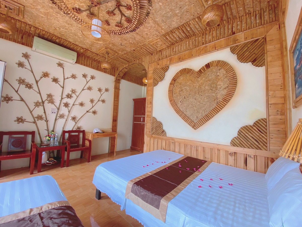 A bedroom featuring two spacious beds adorned with light blue linens and decorative brown throws. The walls have a heart-shaped art piece made of natural materials, and floral patterns are displayed. A small seating area is situated with two wooden chairs and a table in the corner.