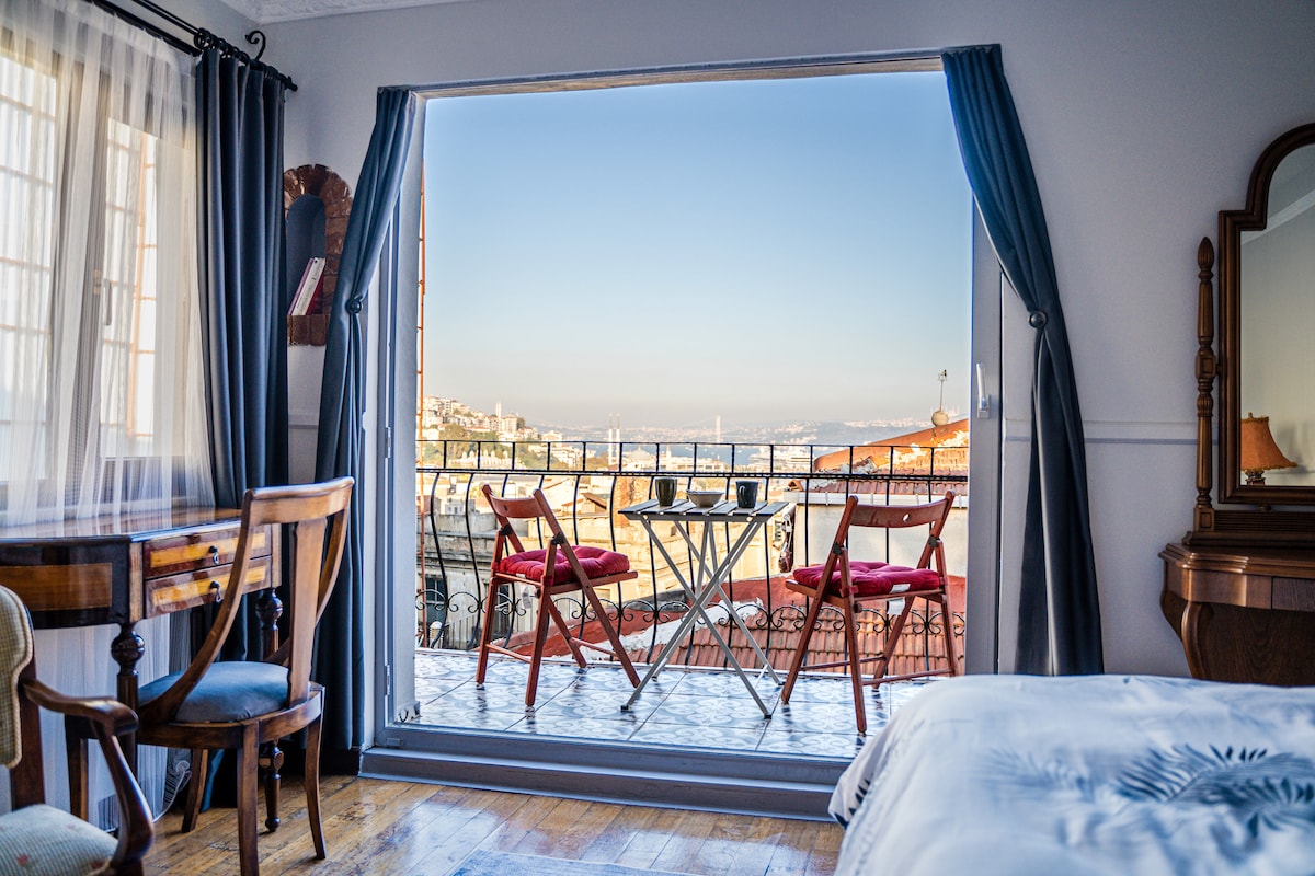 A view from an elegantly appointed room opens to a private balcony with two red chairs and a small table. The spacious interior features wooden flooring, a large window framed by grey curtains, and a wooden desk, enhancing the overall charm of the setting.