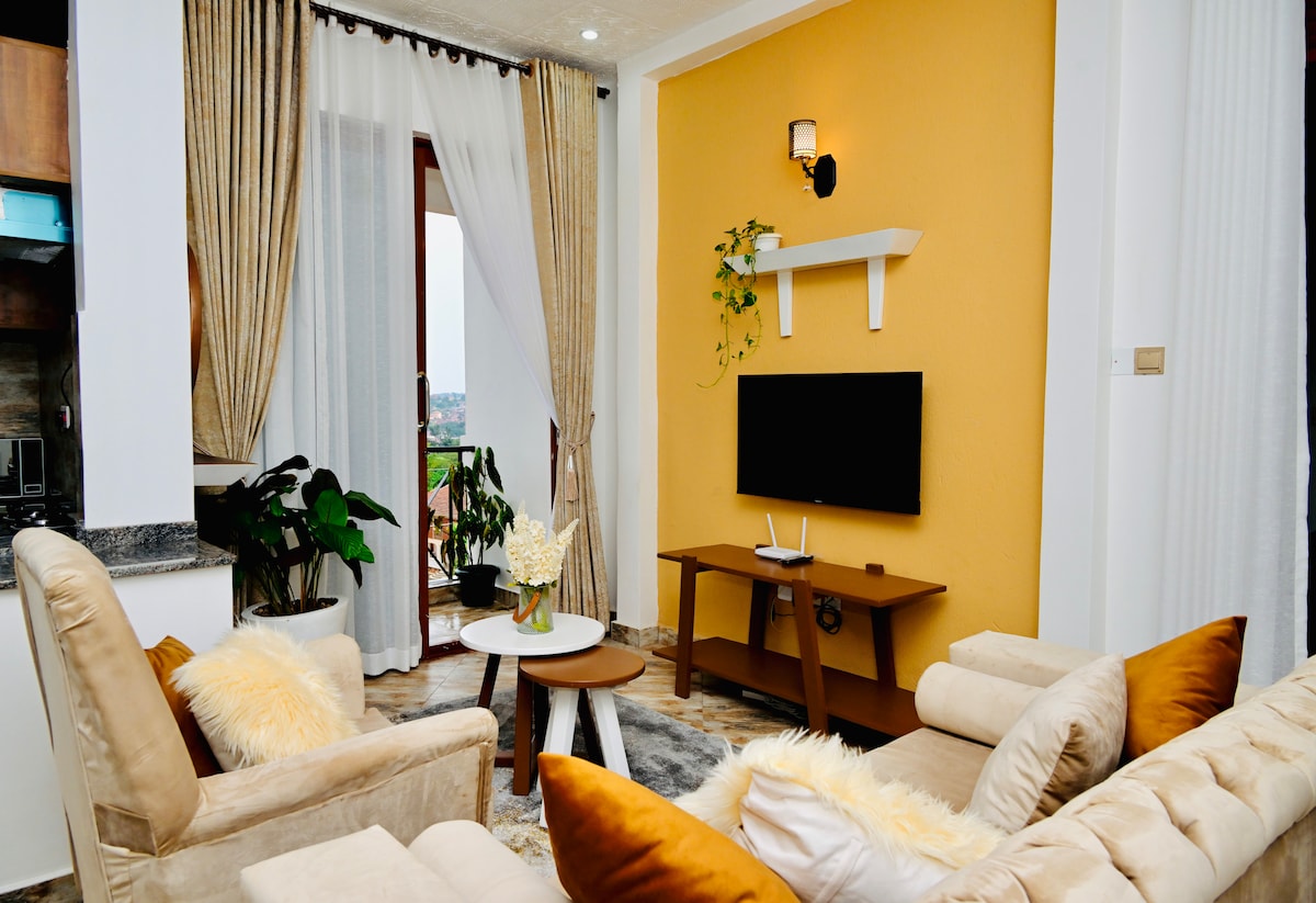 A cozy living area is highlighted by a warm yellow accent wall. Soft, light-colored seating arrangements are complemented by a small round table. Large windows with sheer curtains invite natural light, while indoor plants add a touch of greenery to the space.