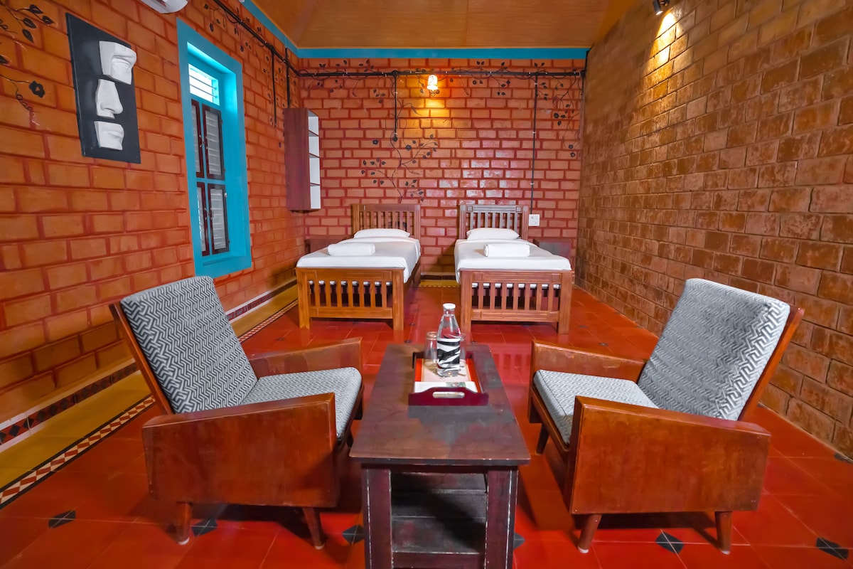 A cozy room is presented with two spacious beds topped with light bedding. Two wooden chairs with patterned cushions are positioned around a low table at the center. Exposed brick walls and colorful accents add warmth, with natural light entering through a window.