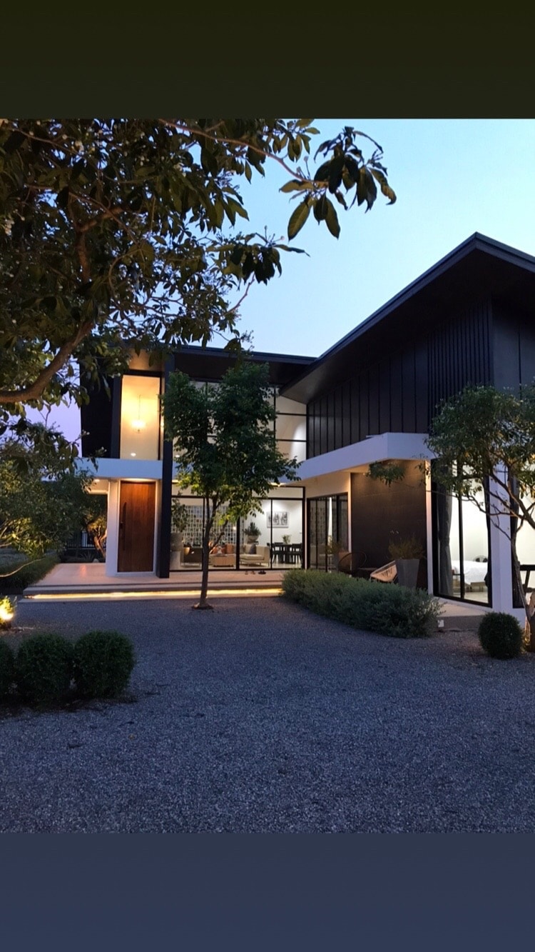 The house features a modern architectural design with large glass windows and a blend of wood and metal elements. The outdoor area is surrounded by softly lit landscaping, including trees and shrubs. A spacious patio is visible, providing a welcoming entryway to the home.