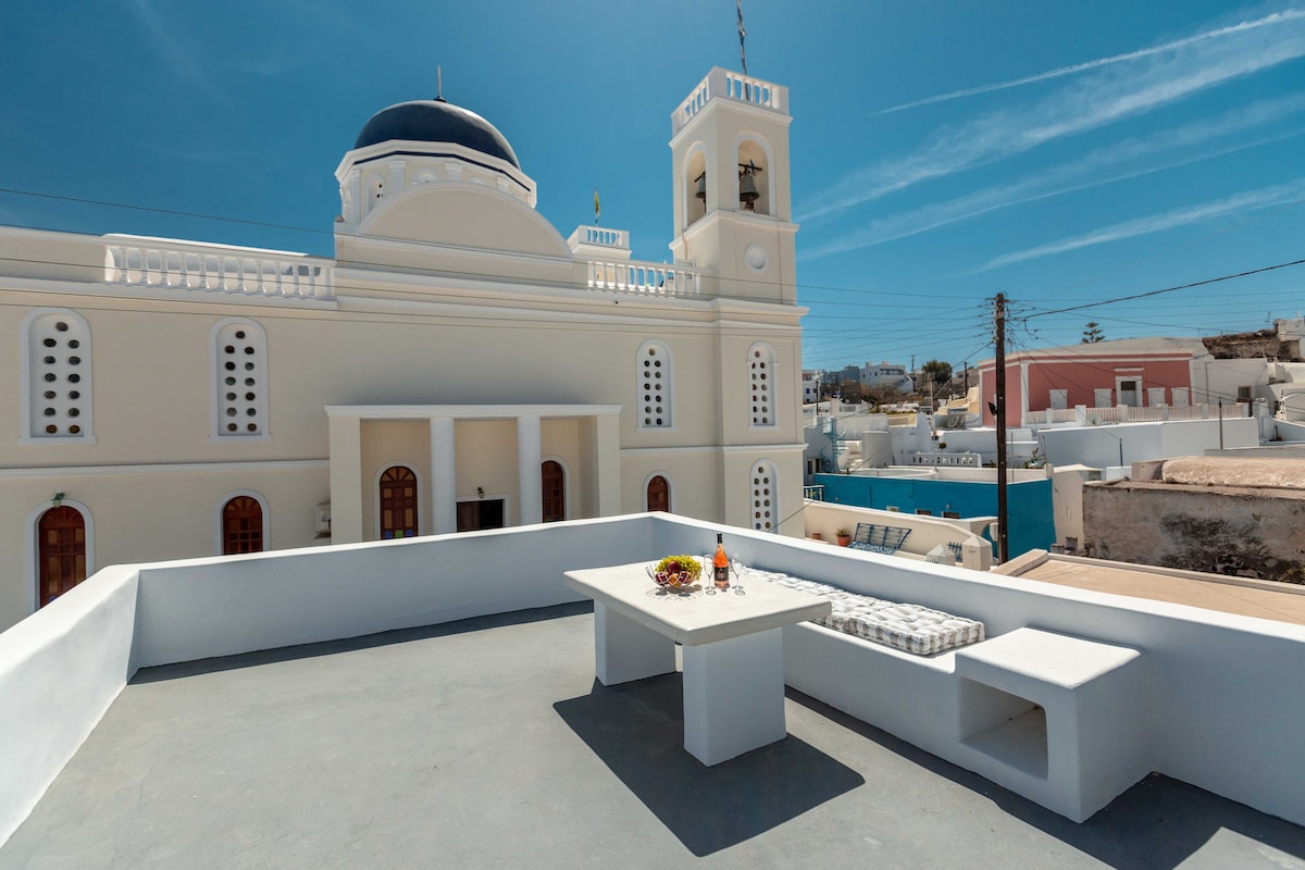 A spacious rooftop terrace features a dining table adorned with a fruit platter and a set of glasses. The scene is complemented by views of a domed church and charming architecture, with blue skies providing a serene backdrop.