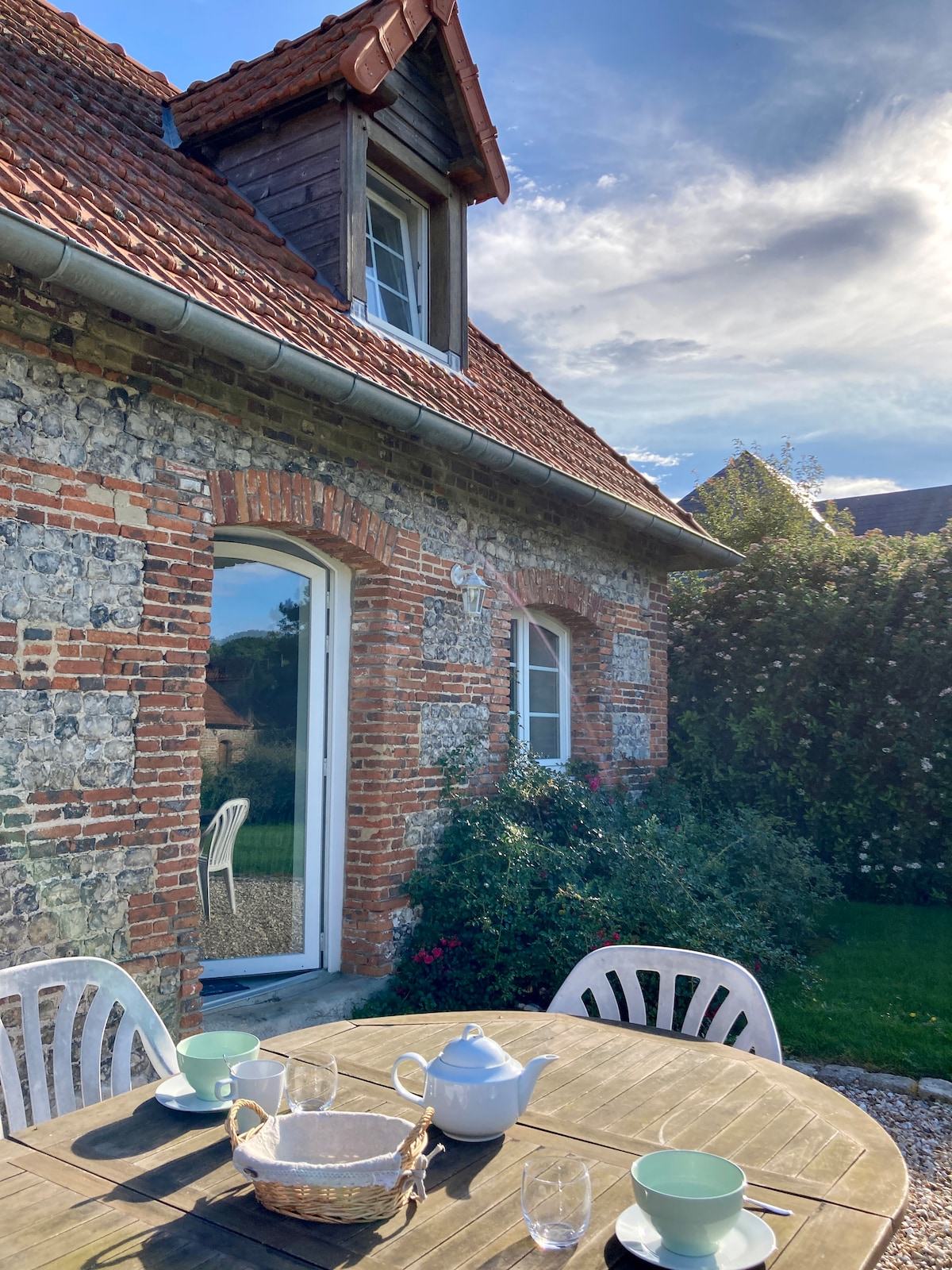 A charming exterior of a stone and brick house features a white door and window, with a neatly maintained garden surrounding it. A wooden table set with tea utensils and glasses sits outside, complemented by white chairs, amidst greenery and a clear sky.