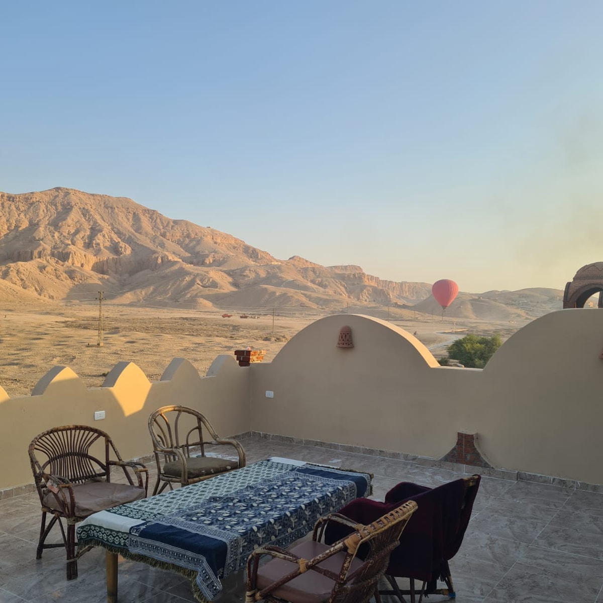 An outdoor terrace is depicted, featuring a table surrounded by four chairs, overlooking expansive arid landscapes. A hot air balloon is seen in the distance against a backdrop of hills, while architectural details of the terrace’s wall add interest to the serene view.