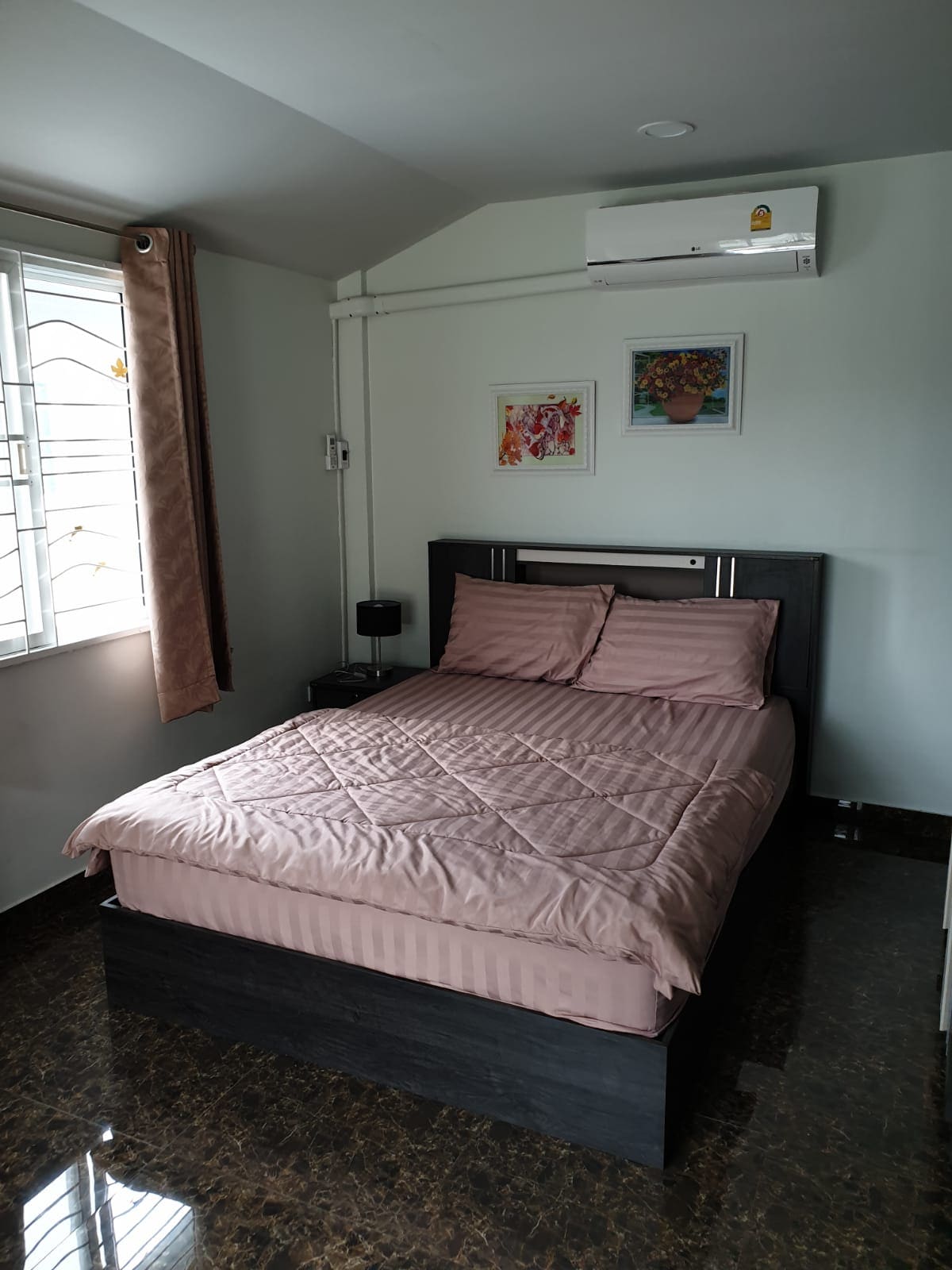 A well-lit sleeping area features a spacious bed dressed in soft pink bedding. Light-colored walls complement the room, while two framed artworks add subtle decoration. A window with sheer curtains allows natural light to filter in, enhancing the serene ambiance.