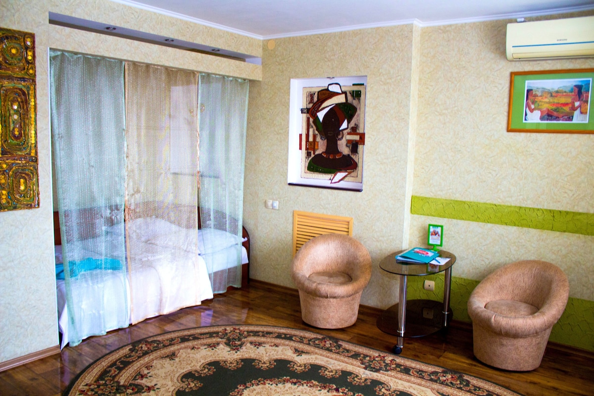 A studio apartment features a sleeping area separated by sheer curtains, revealing a spacious bed. Two rounded armchairs accompany a glass side table on a patterned rug, while a colorful artwork adorns the wall. An air conditioning unit is visible above.