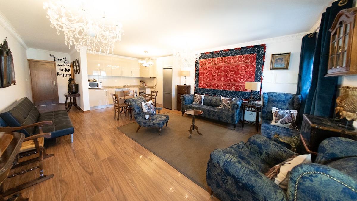 A spacious lounge area features plush seating arrangements, including blue upholstered sofas and armchairs. A decorative rug is centered on the wooden floor, with a coffee table in the middle. Elegant chandeliers provide soft lighting, complementing the adjoining kitchen area in the background.