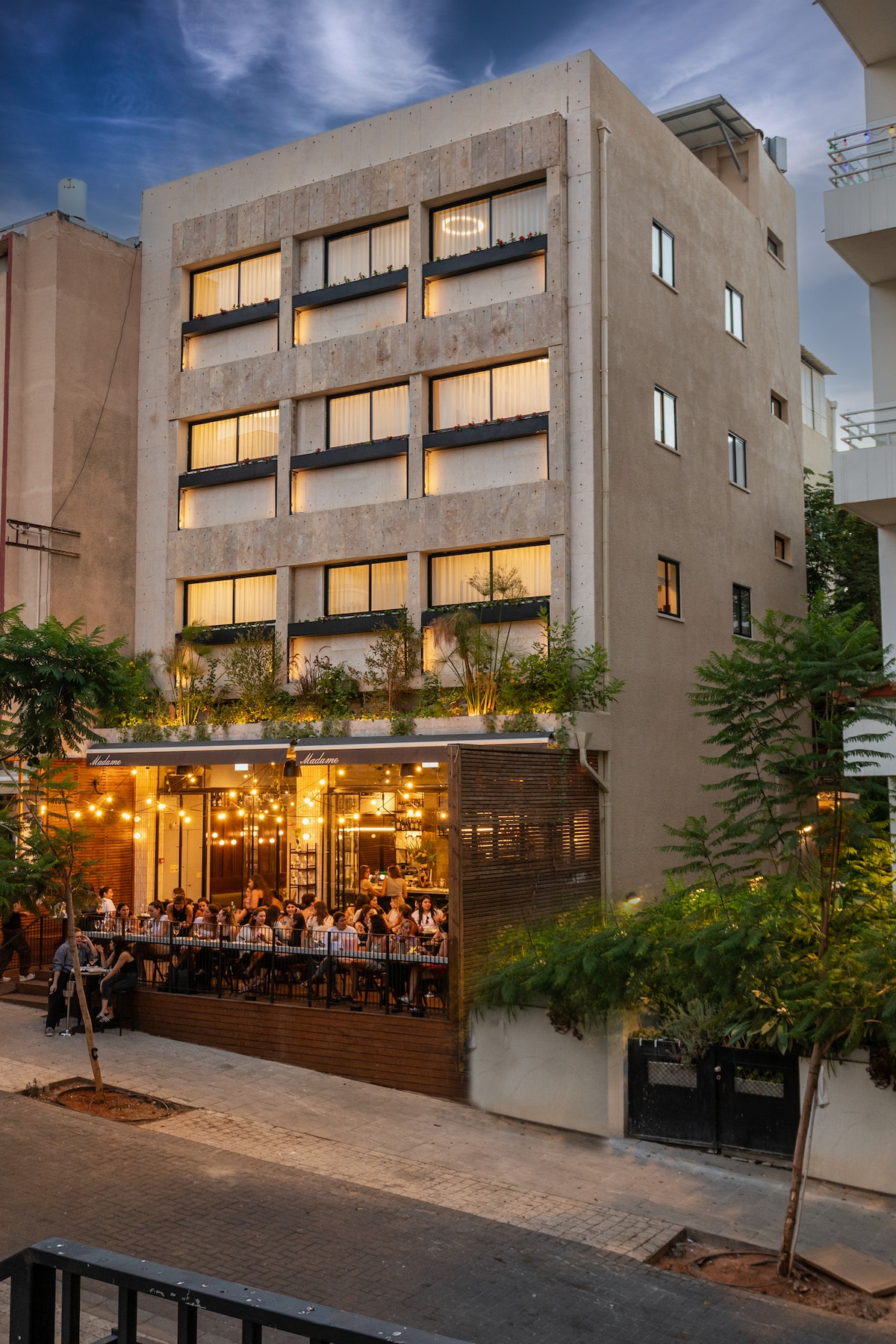 The exterior of a modern building presents a five-story structure with large windows and subtle greenery. A ground-level café features outdoor seating illuminated by decorative lights, creating a welcoming atmosphere. The structure contrasts with nearby buildings, offering a blend of sophistication and comfort.