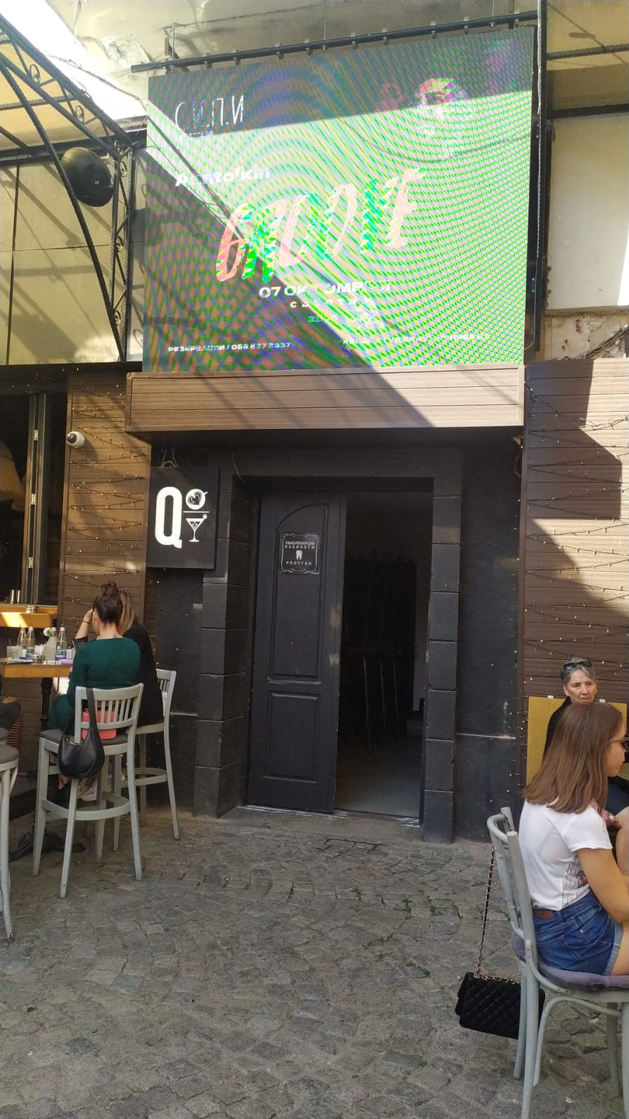 An entrance to a venue is framed by a modern facade, featuring a sleek black door. Above, a vibrant digital sign displays event information. Outdoor seating is visible in the foreground, where patrons are seated at tables, enjoying the lively atmosphere.