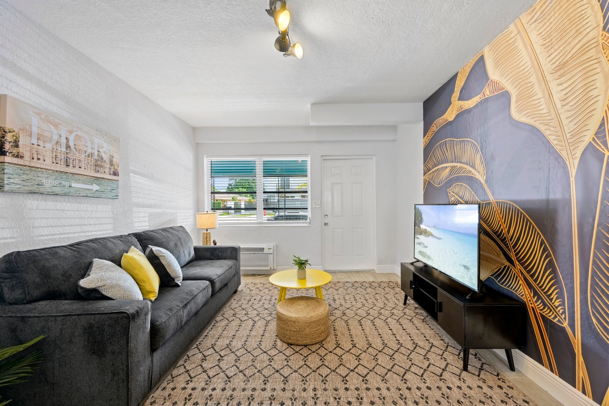 A spacious living area features a dark sofa adorned with colorful pillows, paired with a round yellow coffee table. A large television rests on a sleek black unit, and large windows allow natural light to brighten the room, complemented by modern tropical wall art.
