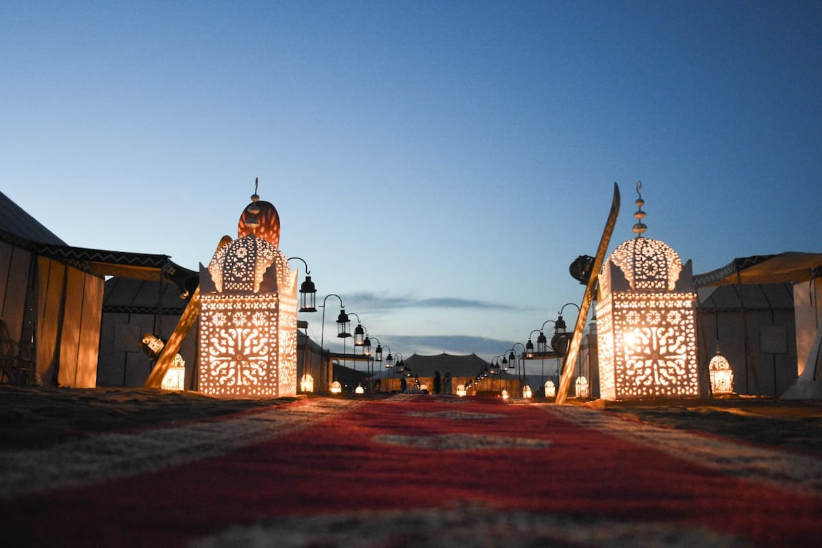 An illuminated pathway leads through the dunes, framed by intricately designed lanterns casting warm light. Decorative structures stand on either side, while a patterned rug guides the way toward a serene desert backdrop under a twilight sky.