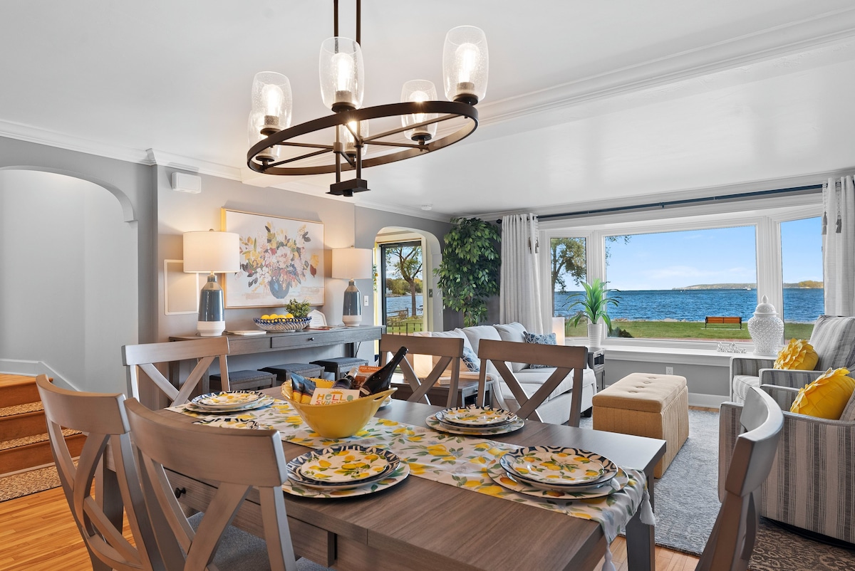 A dining area is equipped with a wooden table set for six, adorned with floral-patterned dishes. Large windows reveal a water view, while a comfortable seating area is arranged nearby. Soft lighting fixtures provide illumination, and decorative elements include plants and artwork.
