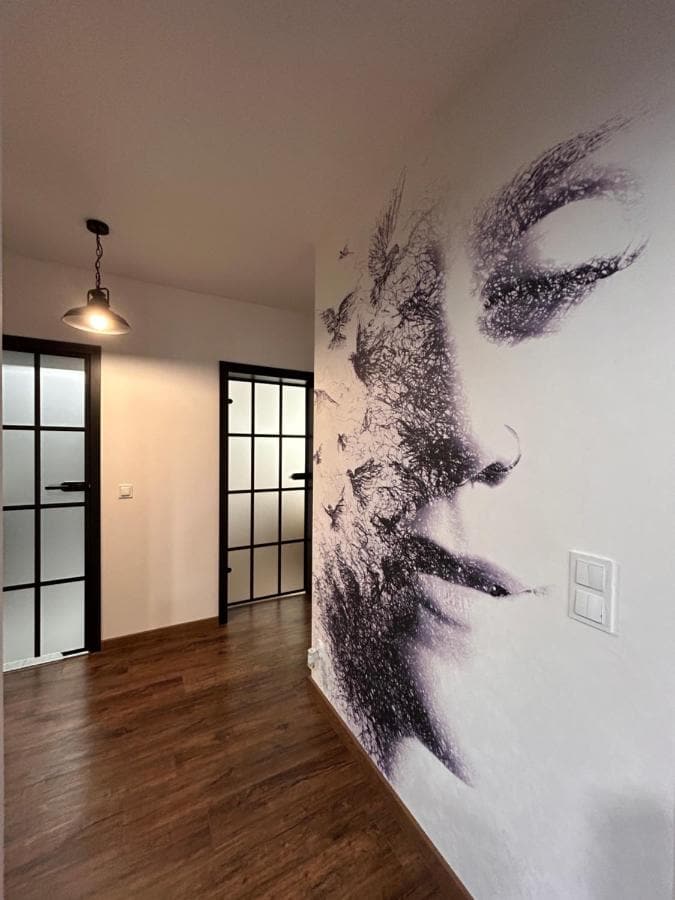 A hallway is presented with contemporary lighting fixtures above. A striking mural of a face blends with artistic elements, creating a unique visual feature. Black-framed glass doors lead to adjacent rooms, complemented by warm wood flooring that enhances the overall ambiance.