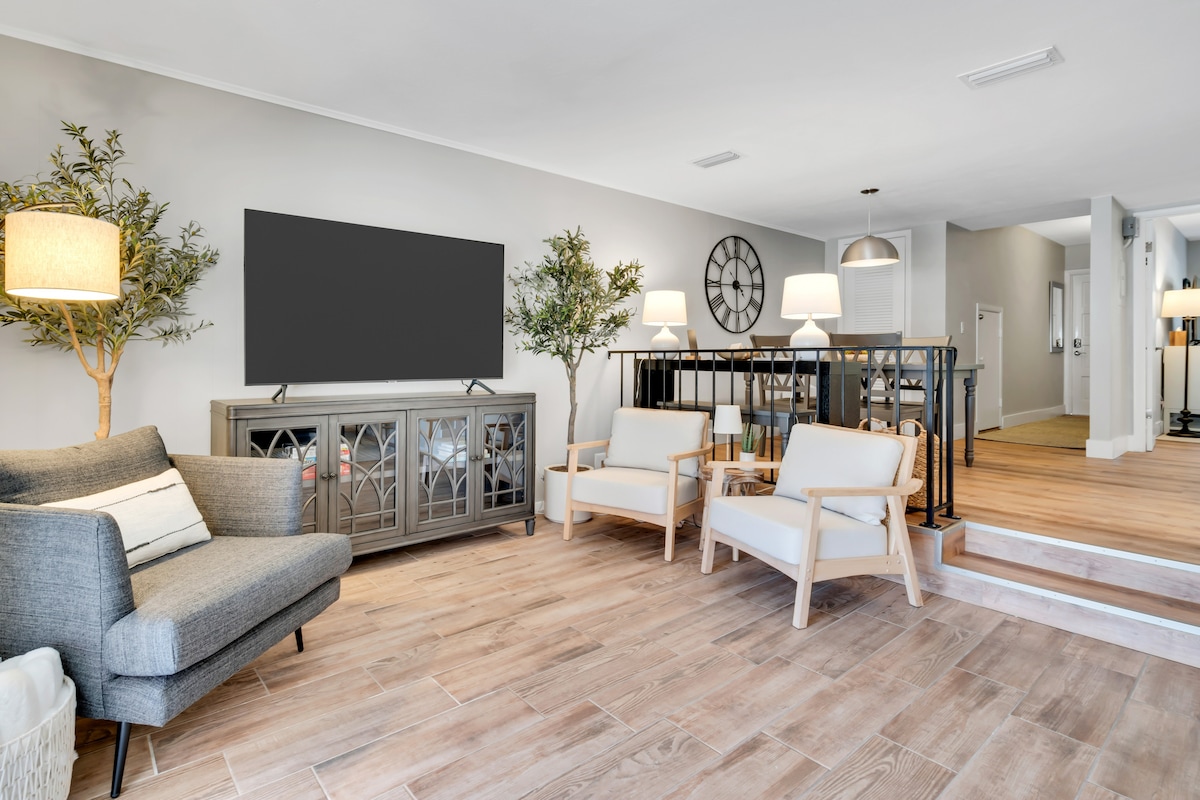 A spacious sitting area features a gray sofa and two white armchairs. A large television is mounted on the wall, complemented by decorative trees and a side lamp. Light wood flooring enhances the bright and open atmosphere.