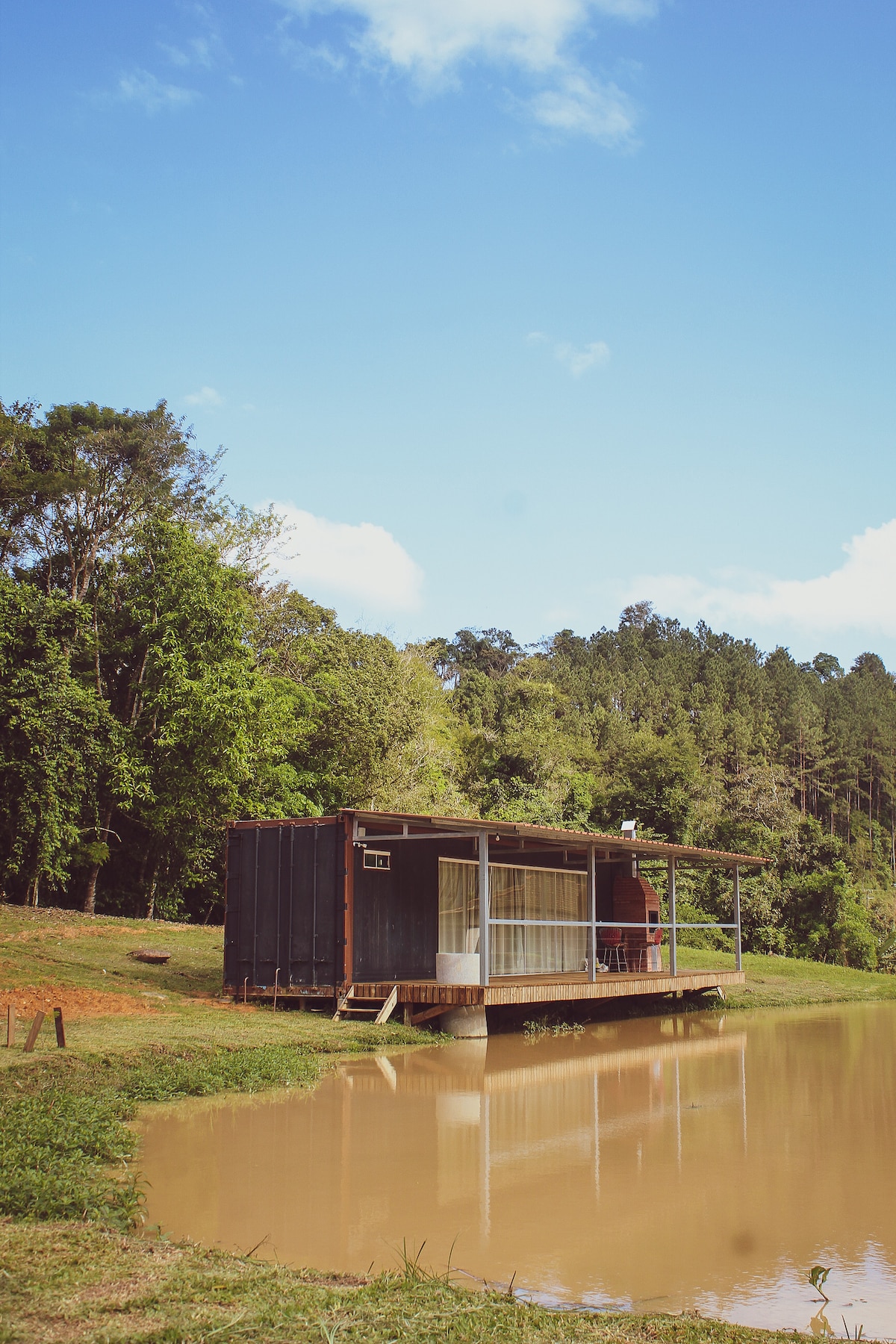 A contemporary container cabin rests beside a tranquil lake, featuring large windows that provide expansive views of the surrounding nature. Lush trees line the background under a clear blue sky, reflecting the serene rural setting.