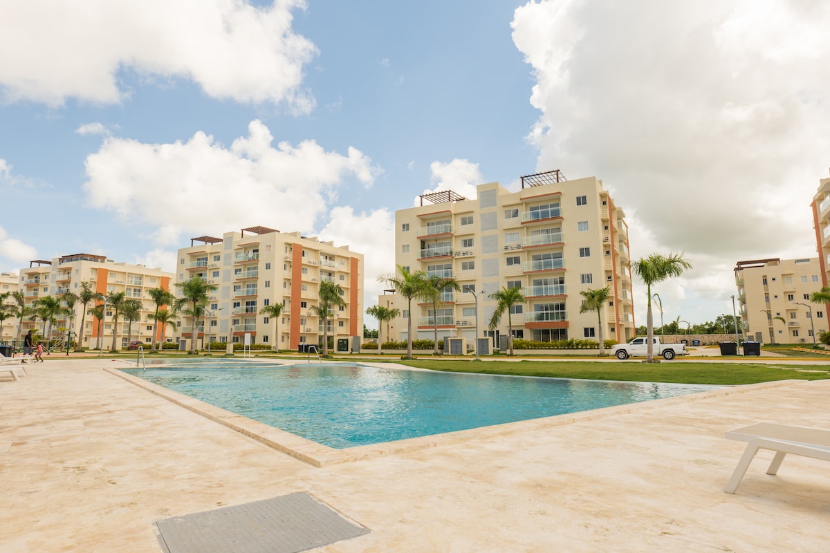 A spacious swimming pool reflects the sky, surrounded by well-maintained landscaping. Modern low-rise buildings with balconies are visible in the background, and palm trees are strategically placed along the pool area, enhancing the serene atmosphere.