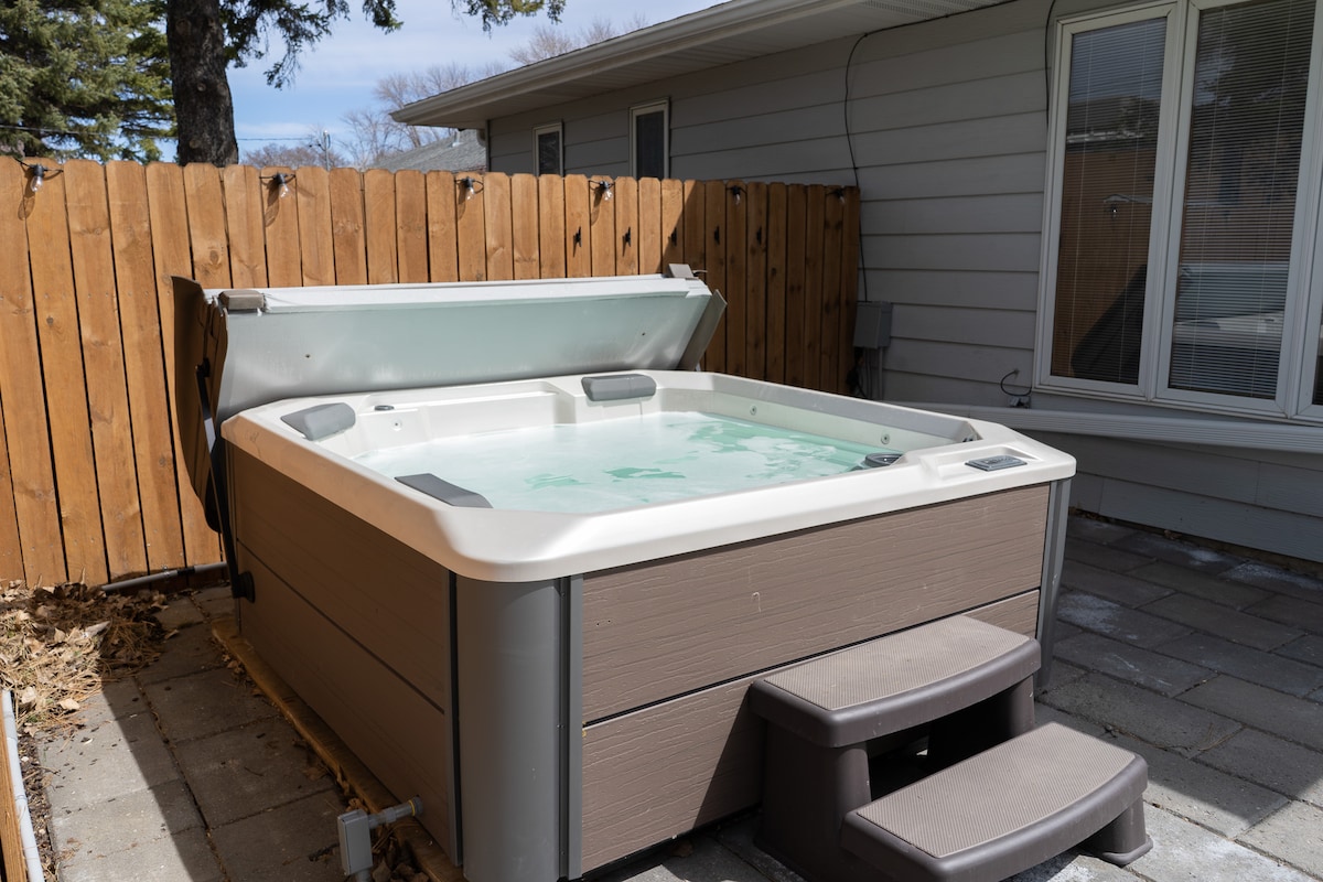 A private hot tub is situated in a fenced outdoor area, surrounded by paving stones. Steps lead up to the hot tub, which features a hinged cover. Natural light illuminates the tub, enhancing the relaxing atmosphere of the outdoor space.