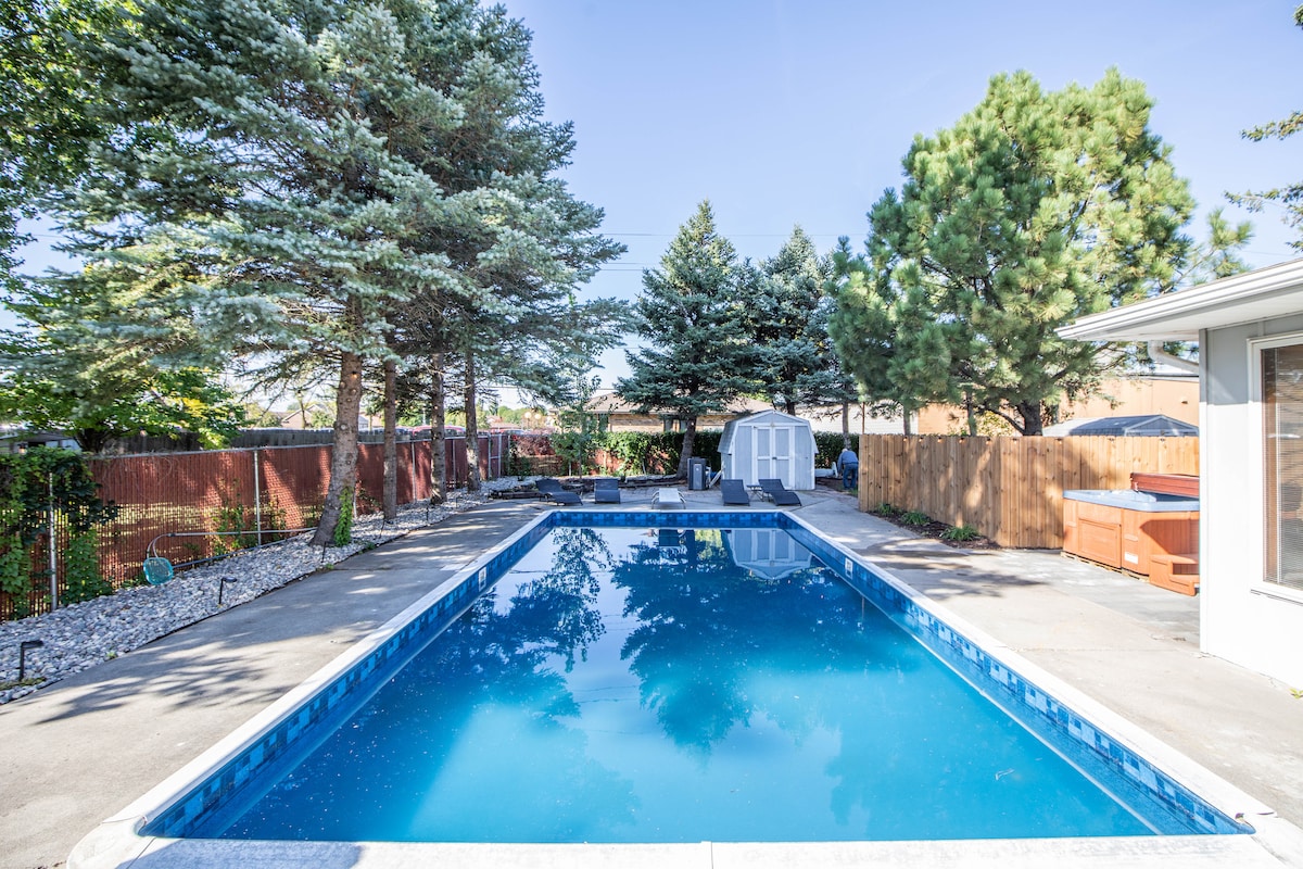 A serene backyard is displayed, featuring a clear in-ground pool surrounded by landscaped greenery and tall trees. A cozy hot tub is positioned nearby, with a small shed visible in the background. The surrounding patio area is spacious, ideal for relaxation.