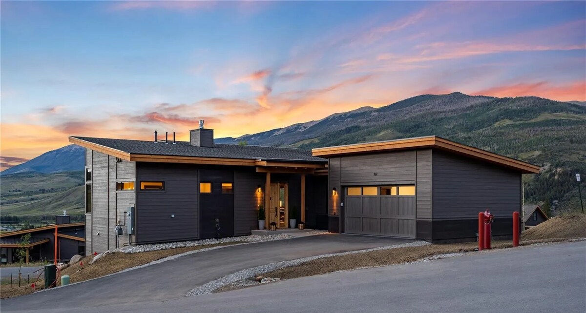A modern two-story vacation house is shown against a colorful sunset, with expansive windows and a two-car garage. The exterior features a combination of dark wood and light paneling, blending seamlessly with the natural landscape of rolling mountains and open fields.
