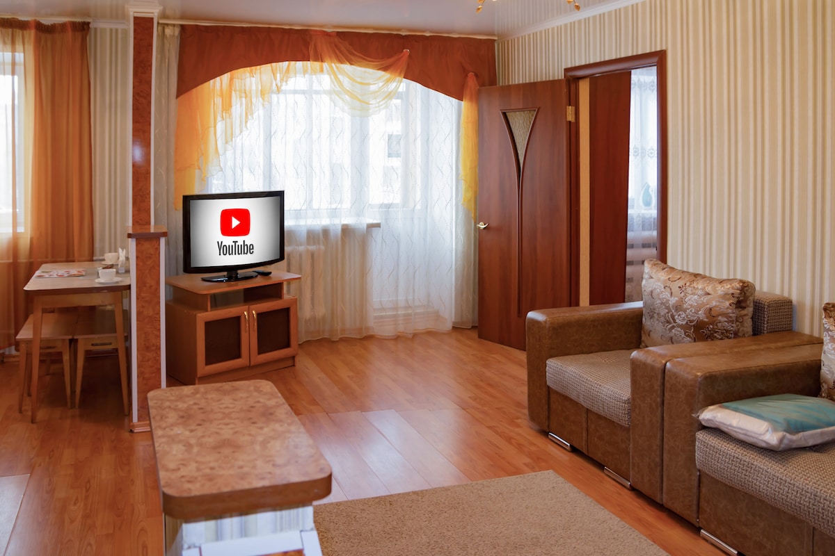 A spacious living area features wood flooring and soft lighting. A comfortable seating arrangement includes two armchairs and a sofa. A television stands on a wooden console, showcasing the YouTube logo. Large windows are dressed in sheer curtains, allowing natural light to brighten the room.
