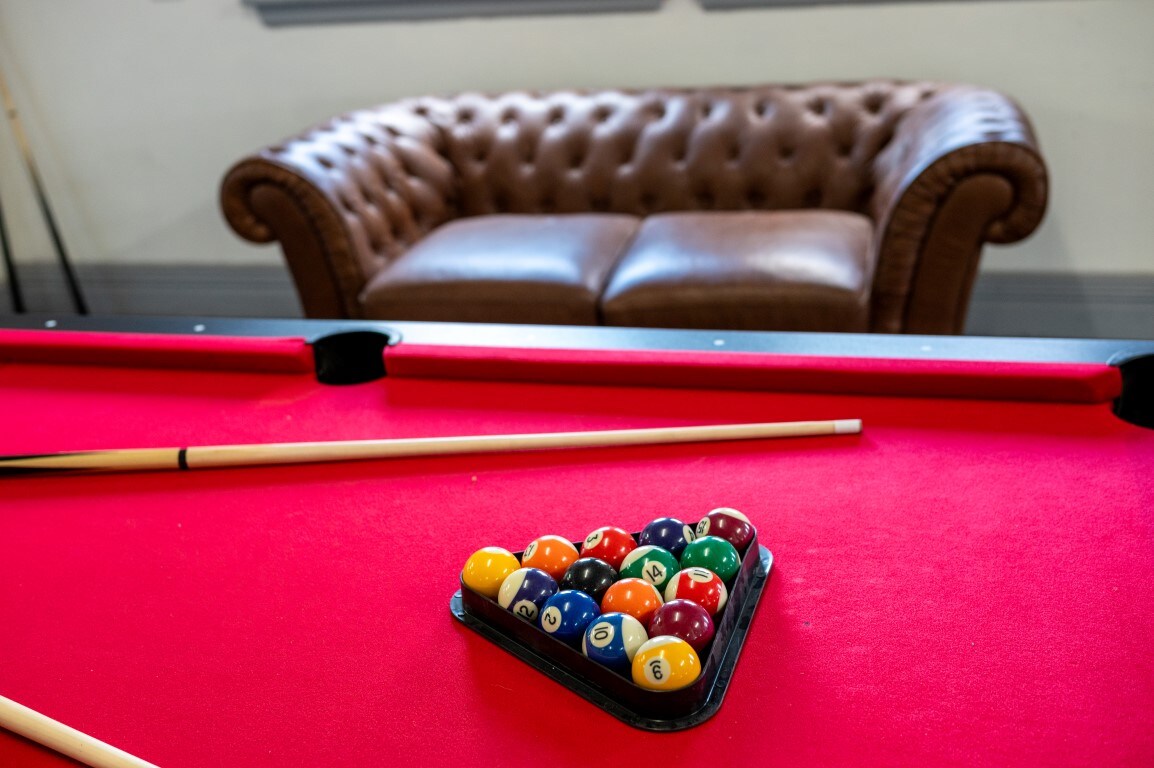 A close view of a billiard table featuring a bright red felt surface, with a triangle rack holding colorful balls positioned at one end. A brown leather chesterfield sofa is visible in the background, providing a comfortable seating area.