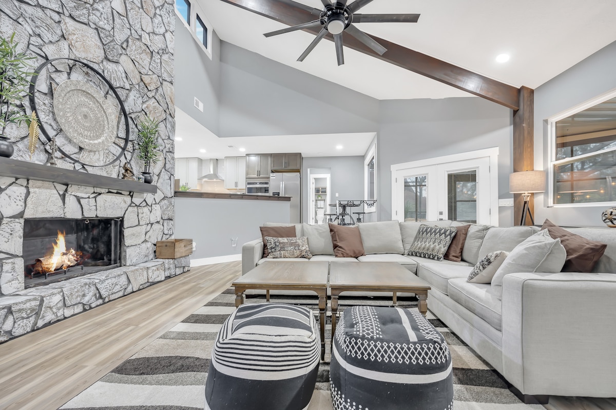 A spacious living area features a stone fireplace, with a large sectional sofa arranged around two wooden coffee tables. Decorative poufs are positioned nearby. Natural light enters through large windows, highlighting the modern design and neutral colour palette.
