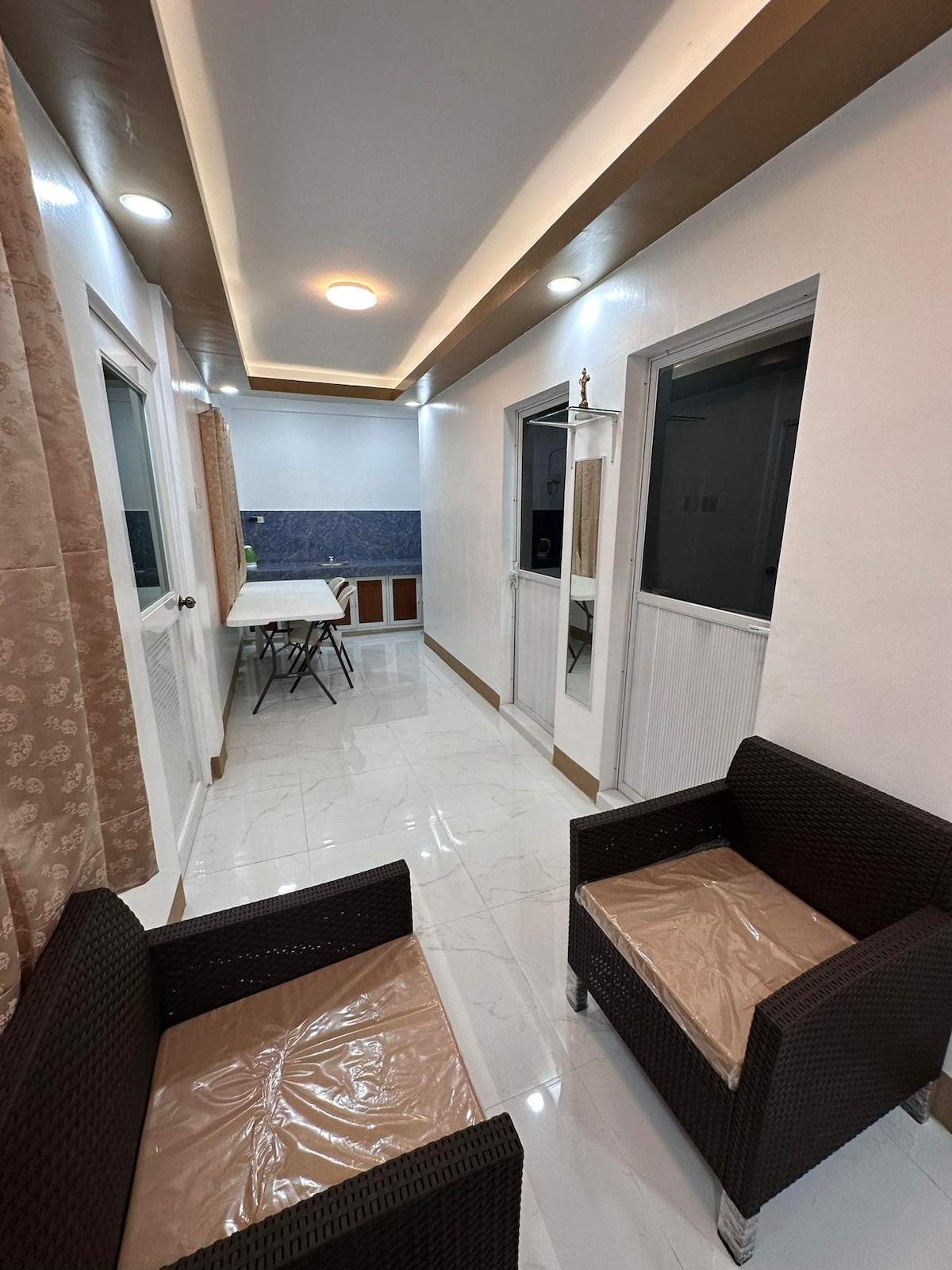 A hallway is shown with glossy white tiles and light-colored walls. Two wicker chairs are positioned at the forefront. To the back, a dining table with chairs is visible, along with large windows allowing natural light to enhance the space.