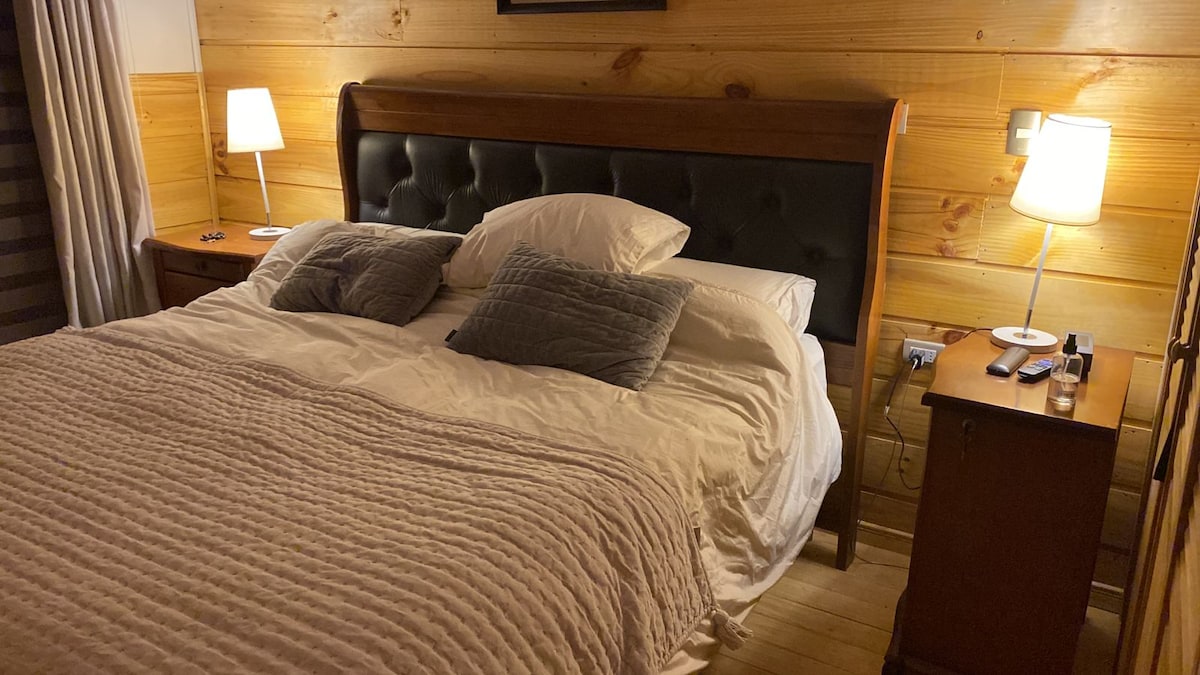 A cozy bedroom features a sleek wooden headboard against warm wooden walls. A spacious bed is dressed in white linens, complemented by two grey decorative pillows. Soft table lamps provide gentle lighting on either side, enhancing the comfortable atmosphere.
