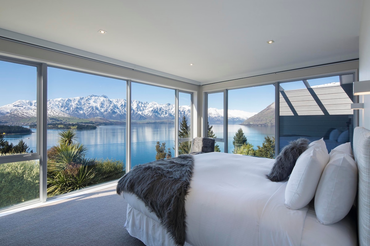 A bright and airy bedroom is featured, with large windows offering panoramic views of mountains and a lake. A spacious bed with decorative pillows is positioned in the center, while soft carpeting adds to the cozy ambiance. Natural light enhances the serene atmosphere.