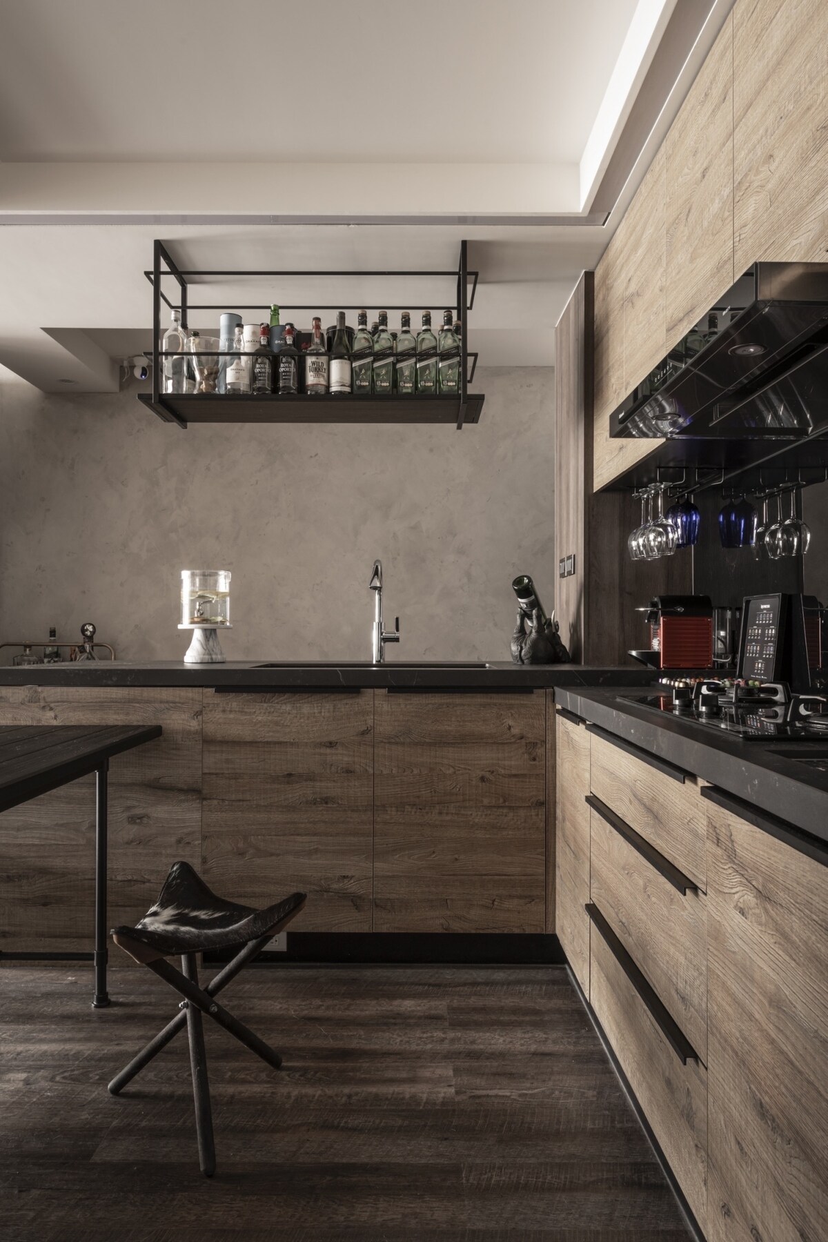 An open kitchen area features wooden cabinetry with a sleek black countertop. A minimalist shelf above displays various bottles. A kitchen workstation is complemented by a unique stool. Modern appliances, including a gas stove, contribute to a functional cooking space.
