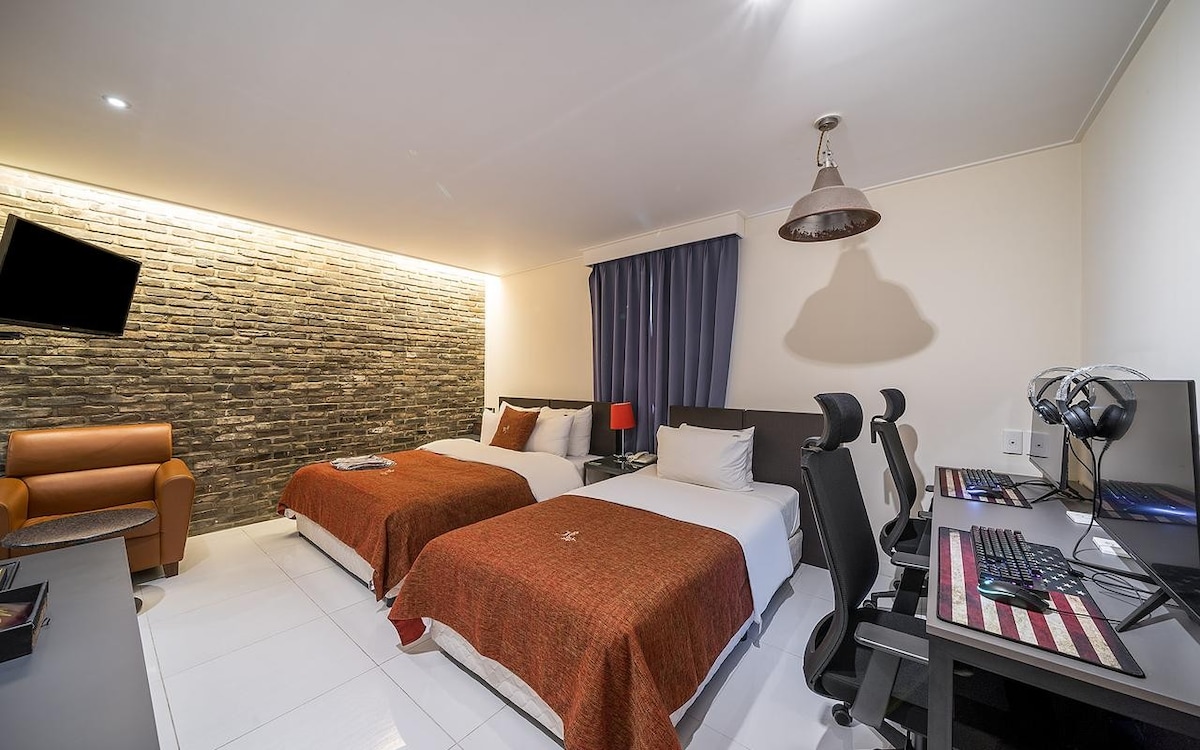 A modern guest room features two spacious beds dressed in warm-toned blankets. A stone accent wall adds texture. A sleek desk with dual computers is positioned nearby, and a brown armchair provides additional seating. Soft lighting enhances the overall ambiance.