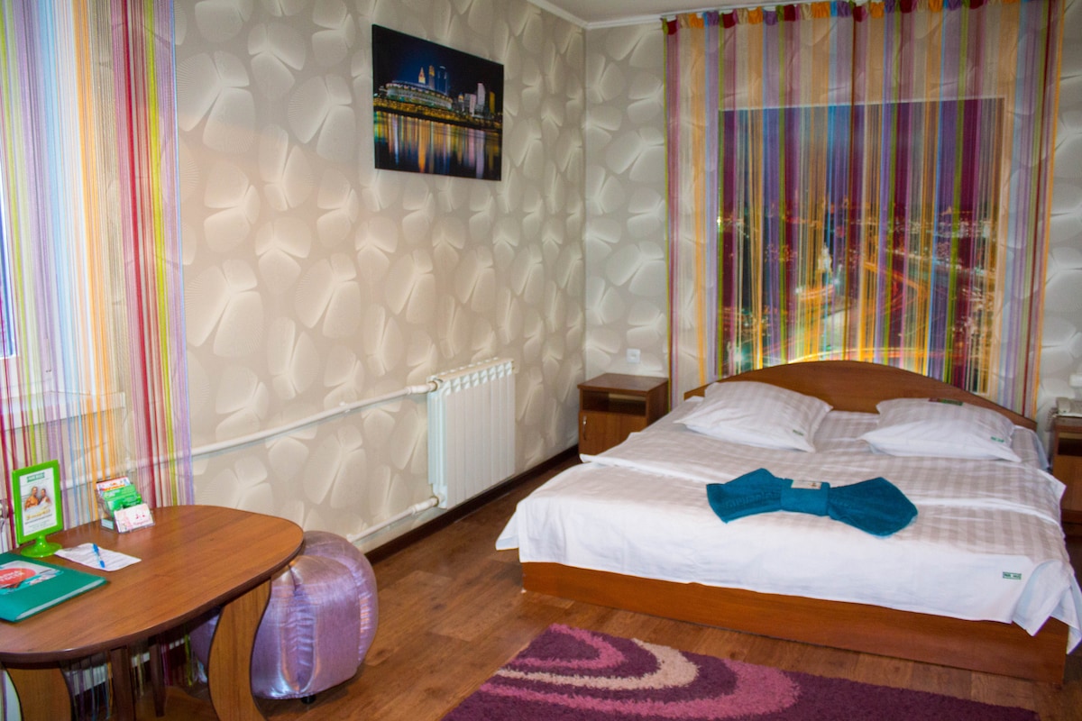 A cozy bedroom is highlighted by a spacious bed dressed in white linens and dark green towels. A round purple ottoman adds a pop of color, while a small wooden table stands beside a colorful area rug. Walls feature a striped curtain and an illuminated cityscape photo.