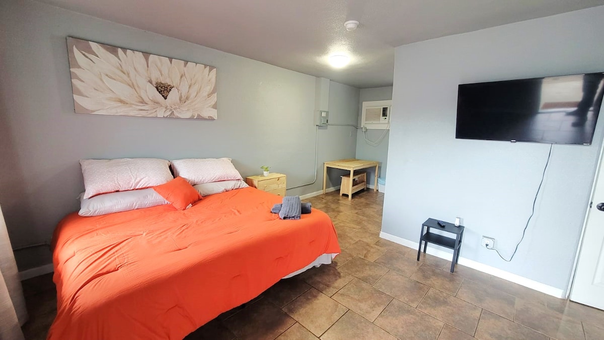 A bright sleeping area features a spacious bed covered with a vibrant orange blanket. A decorative flower artwork hangs above the bed, and a television is mounted on the wall opposite. Natural light enters through a nearby window, illuminating the tiled floor.