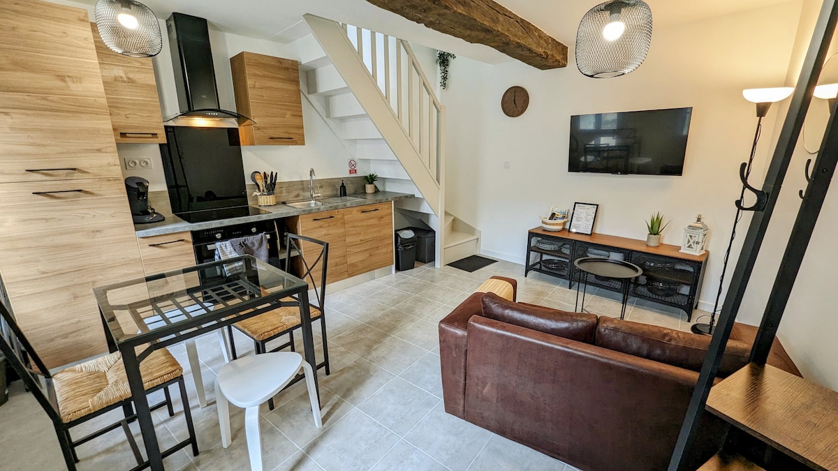 An open-plan living area features a modern kitchen with wooden cabinetry and a dining table accompanied by four chairs. A comfortable brown sofa and coffee table are situated across from a wall-mounted television. Natural light fills the space through a large window, creating a bright atmosphere.