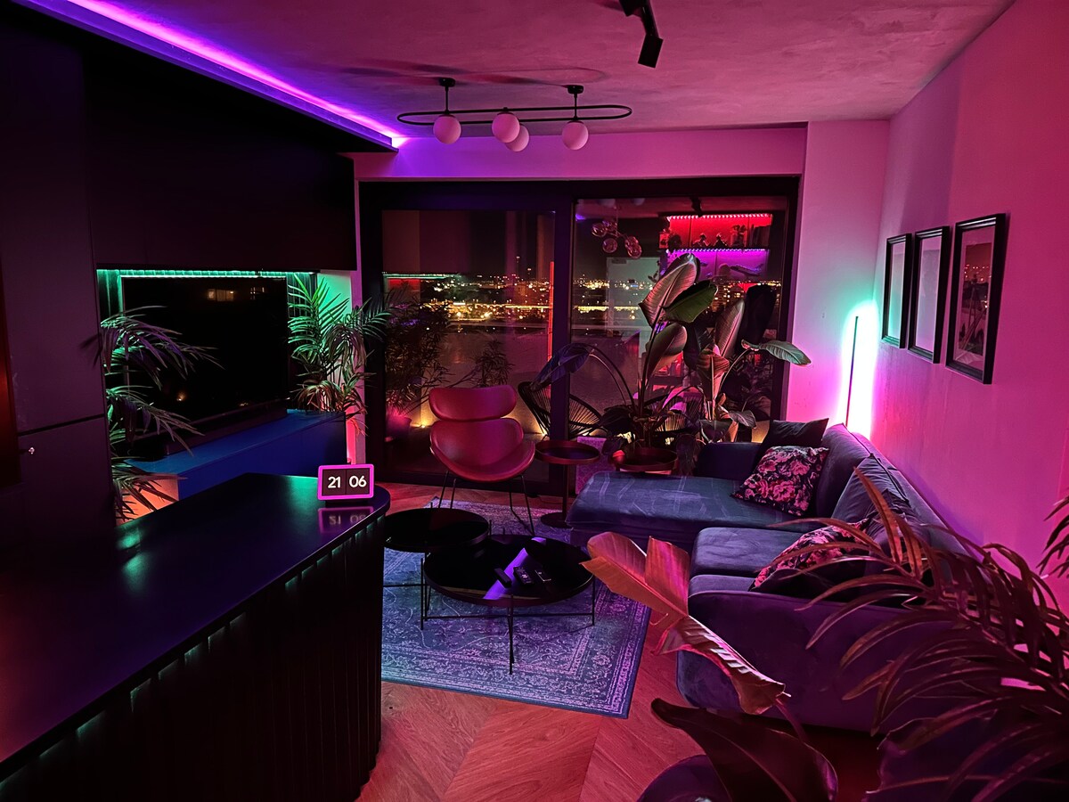 A modern living area is highlighted by ambient lighting in various colors, creating a vibrant atmosphere. A sectional sofa and circular coffee table are positioned in front of a large window, offering expansive views of the city at night. Indoor plants add a refreshing touch.