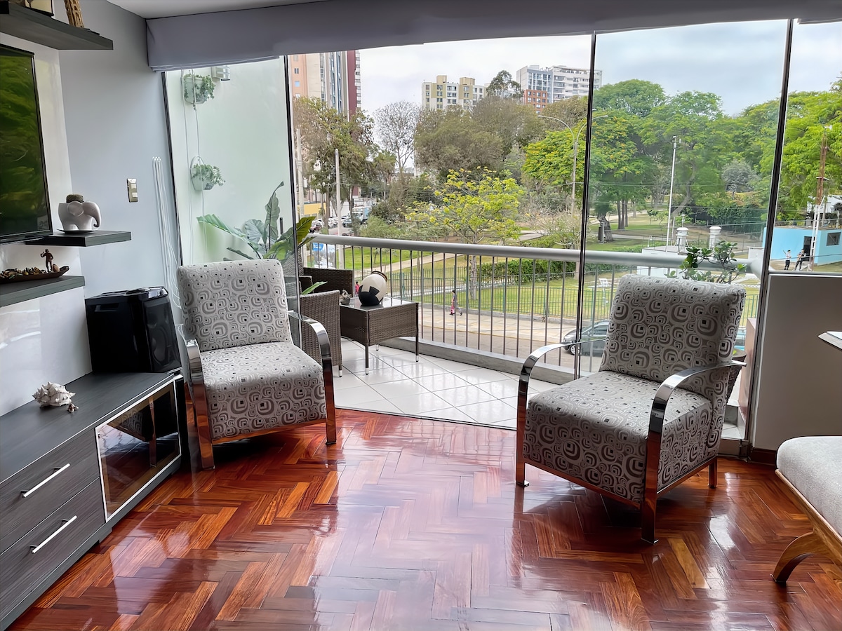 The living area features two comfortable chairs facing large glass doors, offering a view of the greenery in Campo de Marte park. A flat-screen television is placed near the seating area, complemented by a wood-paneled floor that adds warmth to the space.