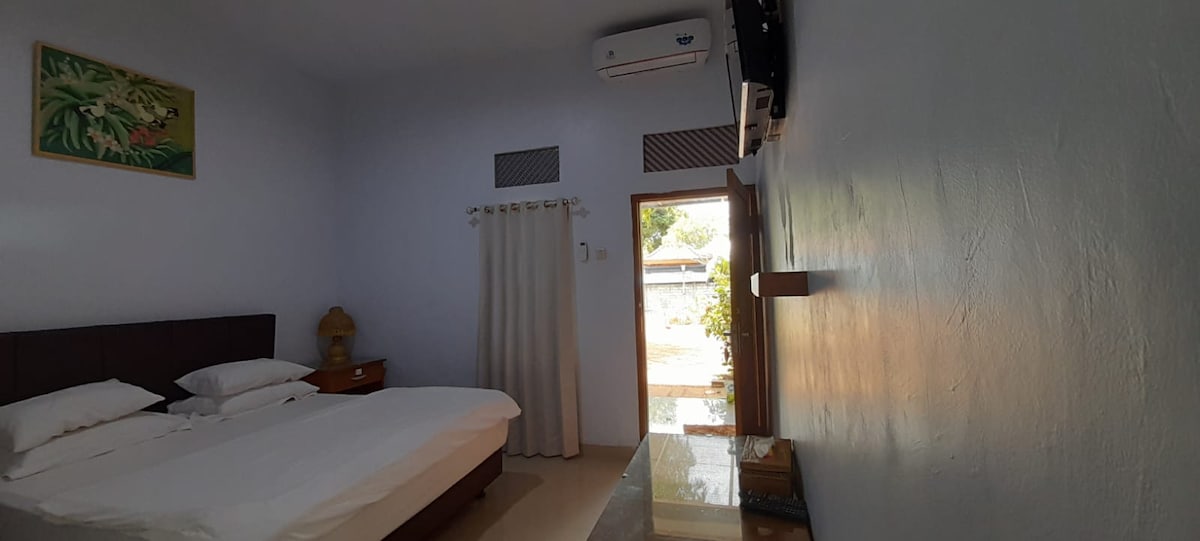 A spacious bedroom features a double bed with white linens and wooden bedside tables. Natural light enters through a door leading to an outdoor area. Air conditioning is installed on the wall, and a television is mounted opposite the bed.