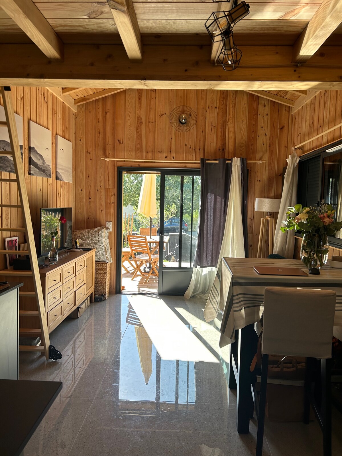 An open interior designed in wood features a cozy dining area with a striped tablecloth. Glass doors lead to a sunlit terrace furnished with outdoor dining. Natural light reflects off the polished floor, while artistic wall decor adds character to the warm space.