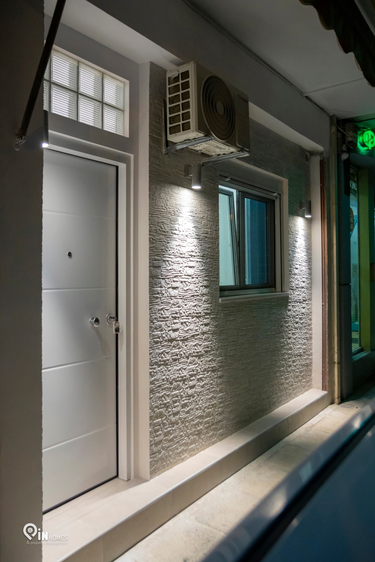 A modern entrance features a sleek, white door with a circular window above. The textured gray wall is complemented by subtle lighting fixtures. An air conditioning unit is mounted above the window, which allows natural light to enter the space.