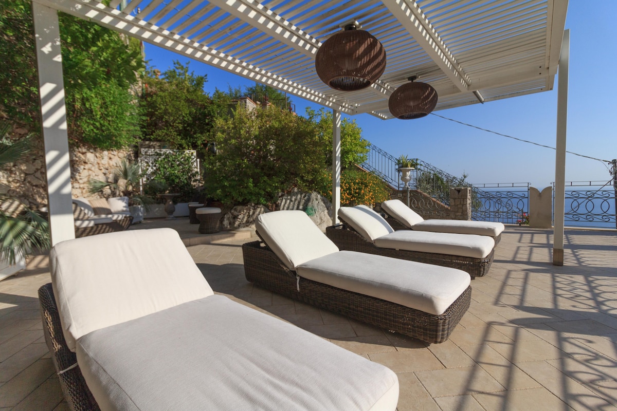 A spacious terrace is equipped with multiple lounge chairs arranged under a shaded pergola. Lush greenery surrounds the area, providing a sense of privacy. The view beyond reveals the sparkling sea, enhancing the relaxation potential of this outdoor space.