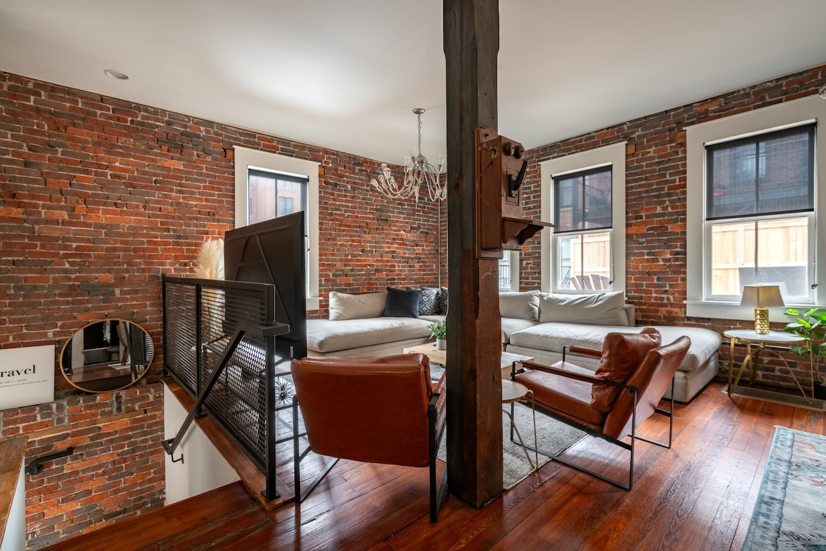 A spacious living area features exposed brick walls and tall ceilings. Large windows allow natural light to illuminate the space, highlighting a mix of comfortable seating and decorative elements. A wooden beam adds architectural interest, while a stylish chandelier provides a focal point above.