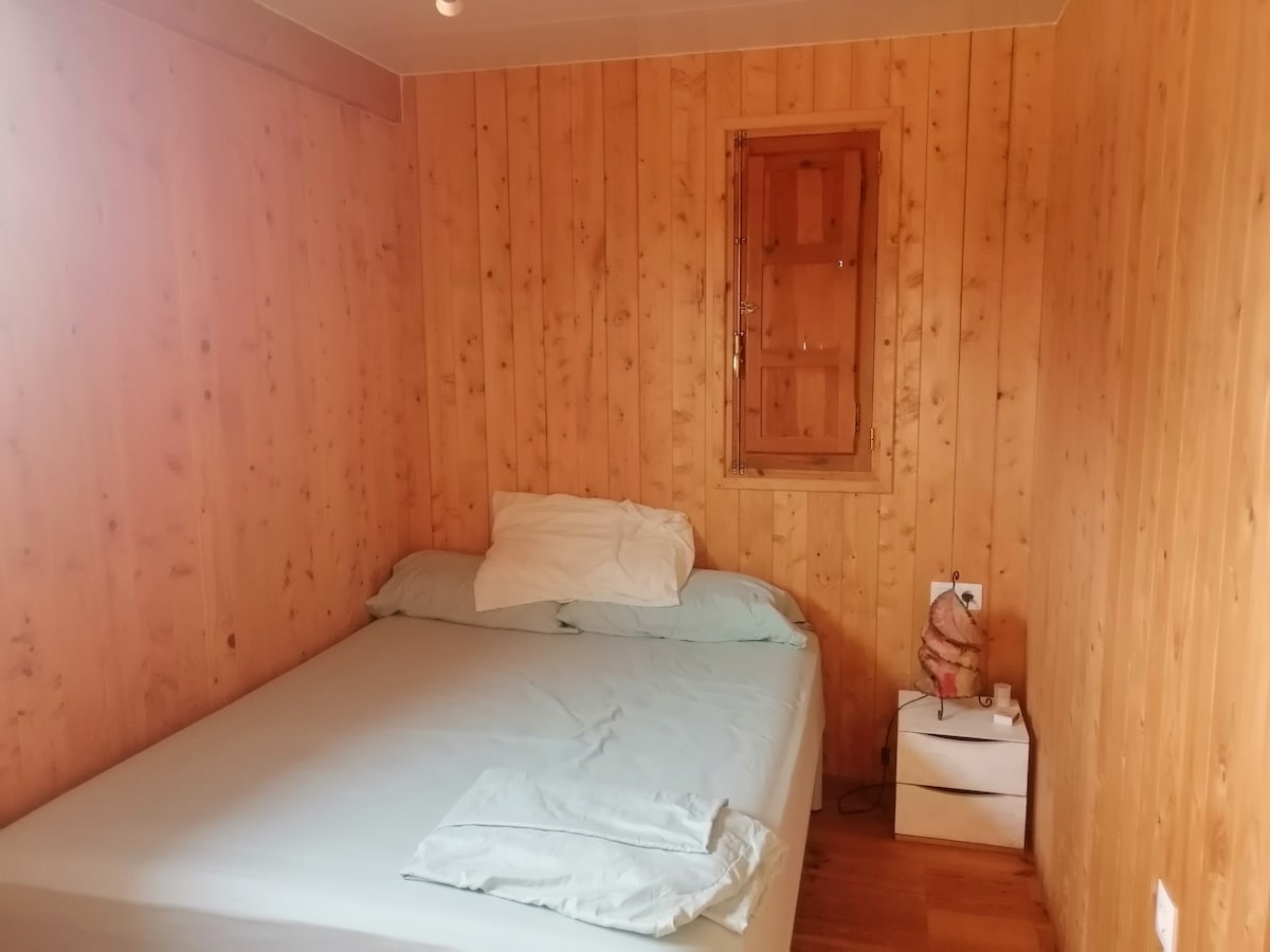 A cozy sleeping area features wooden walls, creating a warm atmosphere. A spacious bed is dressed with light-colored linens, and a few pillows are casually arranged. A small bedside table holds a lamp, while a narrow window provides natural light.