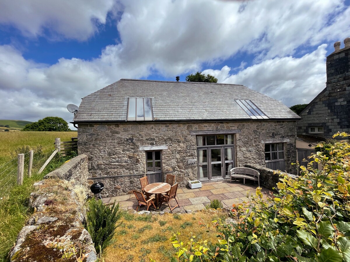 The exterior of a stone cottage is displayed under a partly cloudy sky. A paved patio area features a round dining table surrounded by four wooden chairs, with a charcoal grill nearby. Lush greenery and a grassy field are visible in the background.