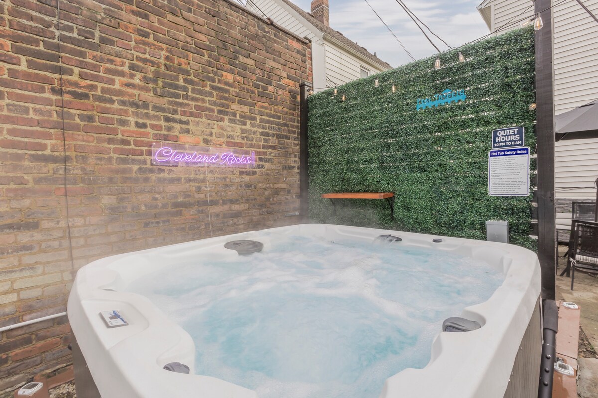 A hot tub with bubbling water is positioned against a textured brick wall, accompanied by a green privacy screen. Soft lighting creates a relaxed environment, while the neon sign displays 'Cleveland Rocks.' Seating options are available nearby for guests to enjoy the space.