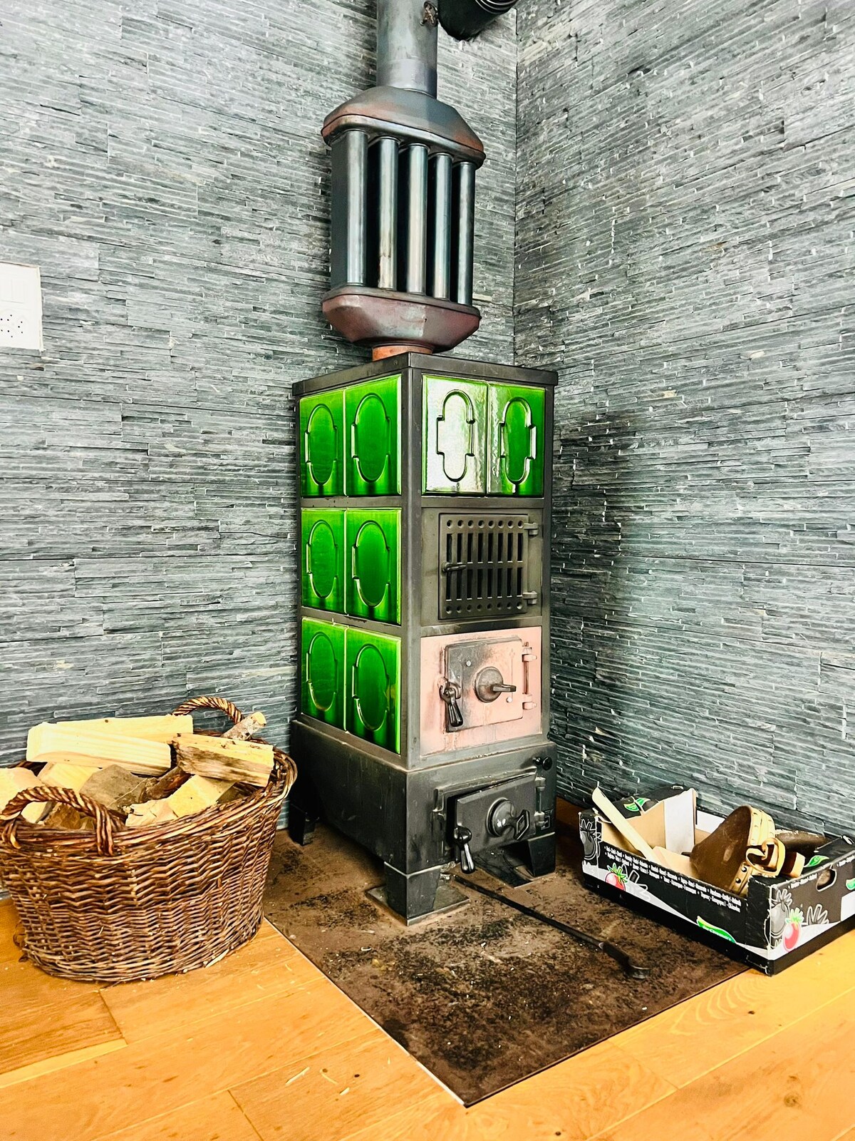 A decorative wood stove features green ceramic tiles and an ornate design, situated against a textured stone wall. A wicker basket filled with firewood is placed beside the stove, contributing to a cozy ambiance. A small box for additional storage is visible nearby.