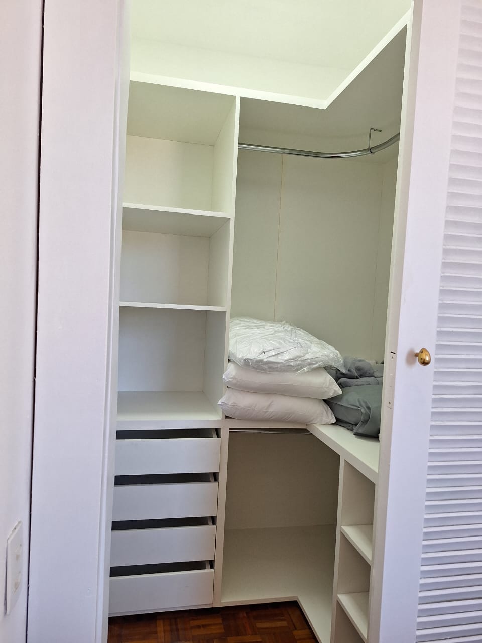 A well-organized closet features built-in shelves and drawers. Freshly laundered bedding is neatly stacked on one side. The open layout allows for easy access to storage, with a hanging rod positioned above the shelves.