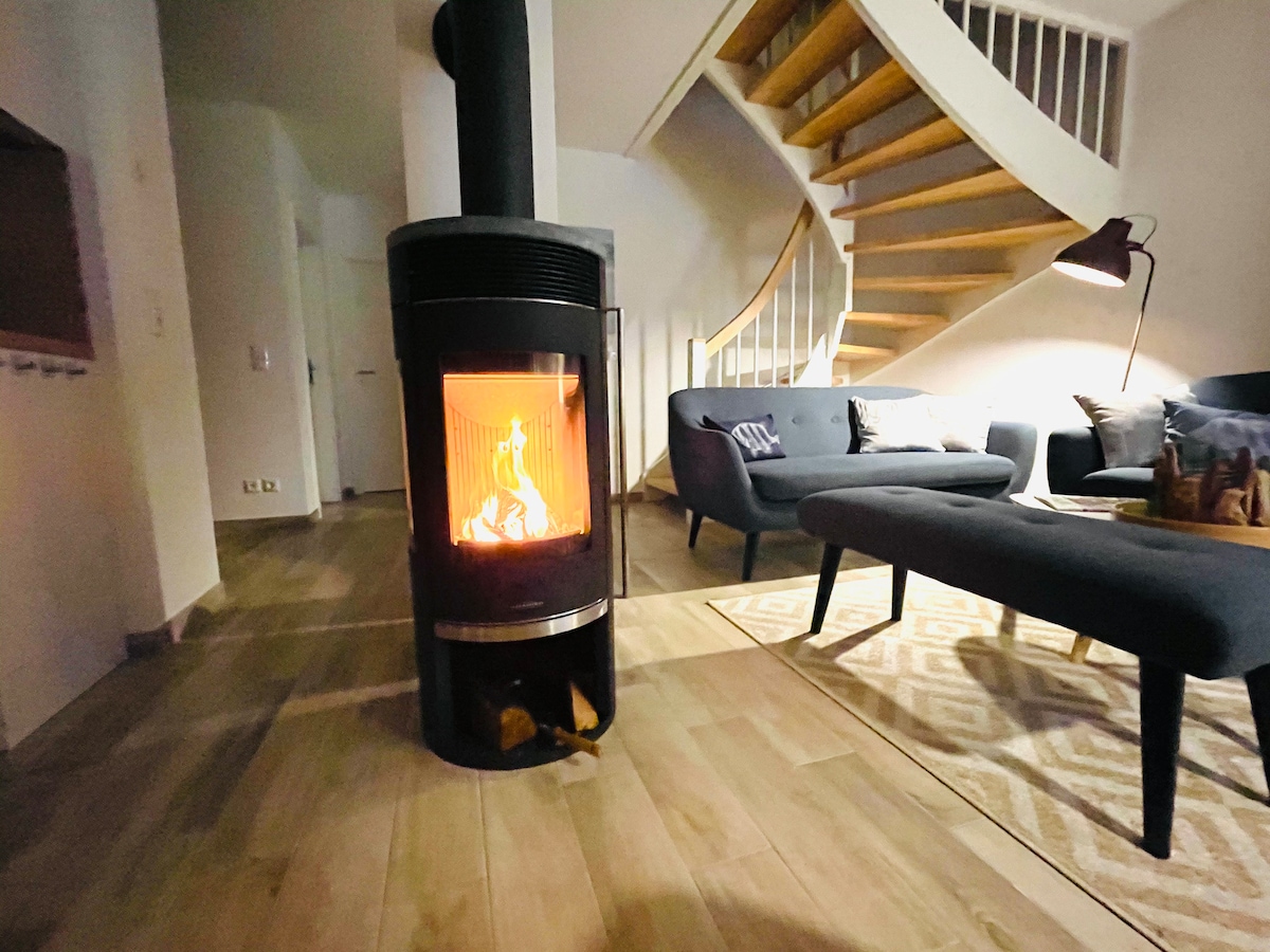 A modern wood stove is centrally located, providing warmth to the room. Soft light reflects off the light wooden flooring. An elegantly curved staircase is visible in the background, complementing the comfortable seating area furnished with dark couches and a patterned rug.