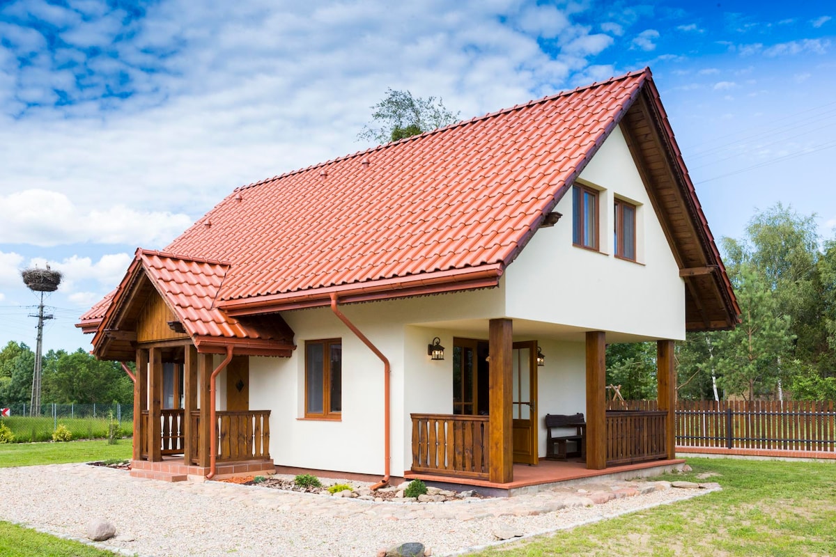 A charming two-story house is displayed, featuring a red tiled roof and cream-colored exterior. Wooden accents frame the entrance and windows, while a gravel path leads up to the porch. Lush greenery surrounds the property, enhancing its serene environment.