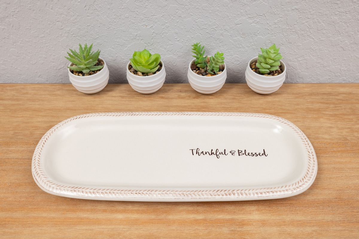 A rectangular serving platter rests on a wooden surface, featuring the phrase 'Thankful & Blessed' in elegant script. Behind the platter, four small potted succulents add a touch of greenery, each in a white ceramic pot with a subtle texture.